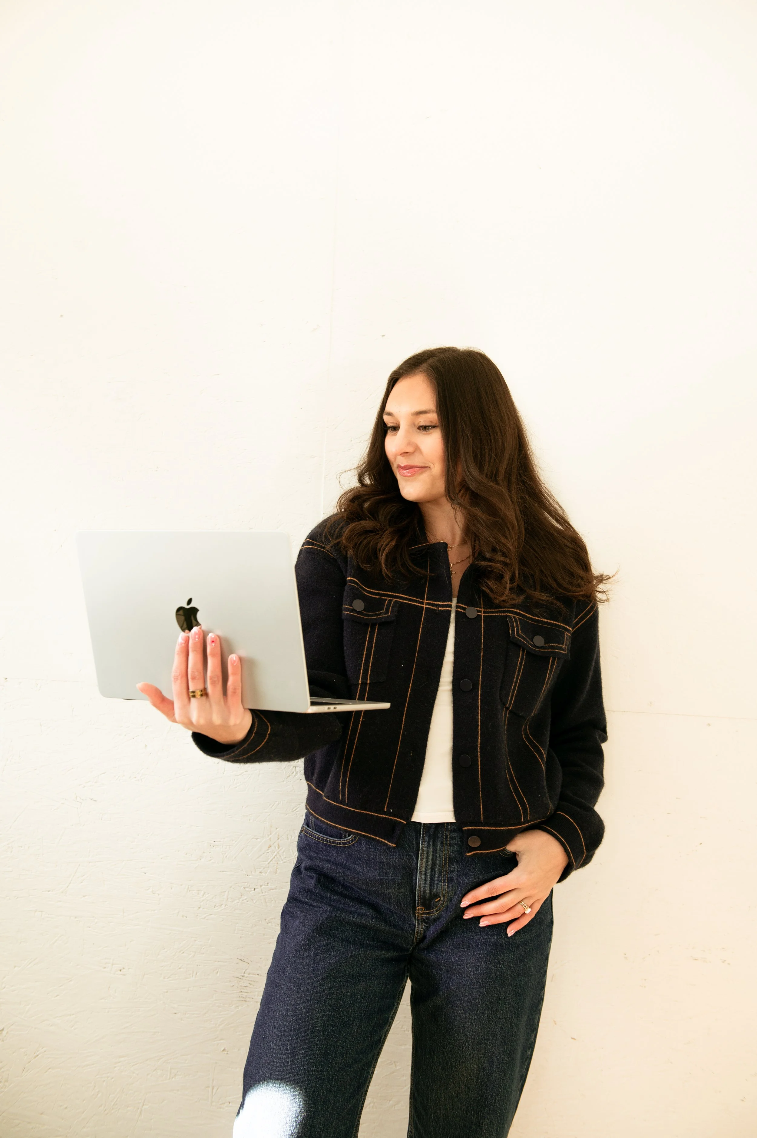 A woman with long brown hair wearing a denim jacket and dark jeans, holding a silver MacBook laptop in her left hand, standing against a plain white wall.