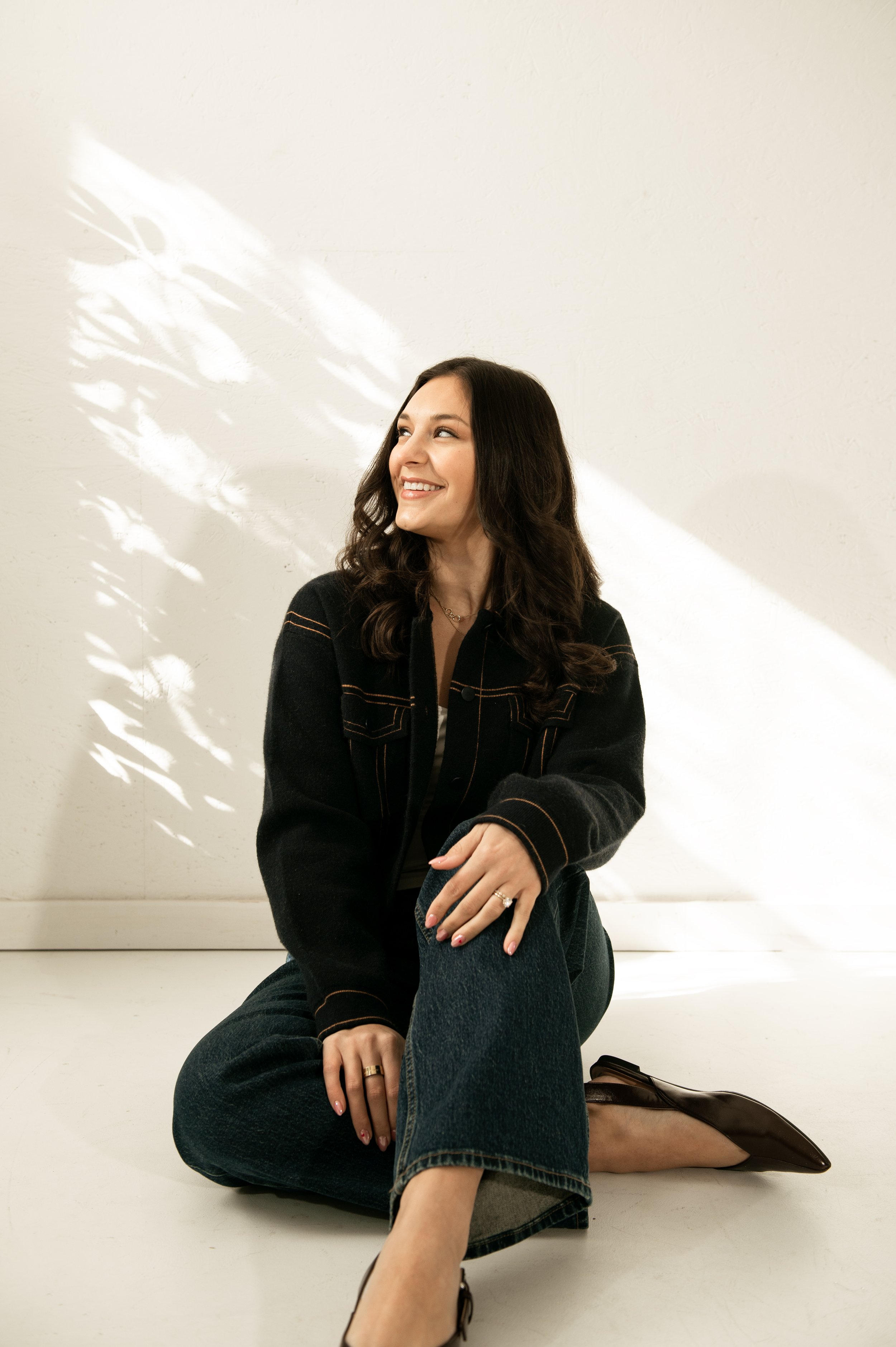 A woman with long dark hair smiling and looking to her left, sitting on the floor with one knee up, wearing a black jacket, jeans, and brown shoes, in front of a plain light-colored wall with sunlight and shadow patterns.
