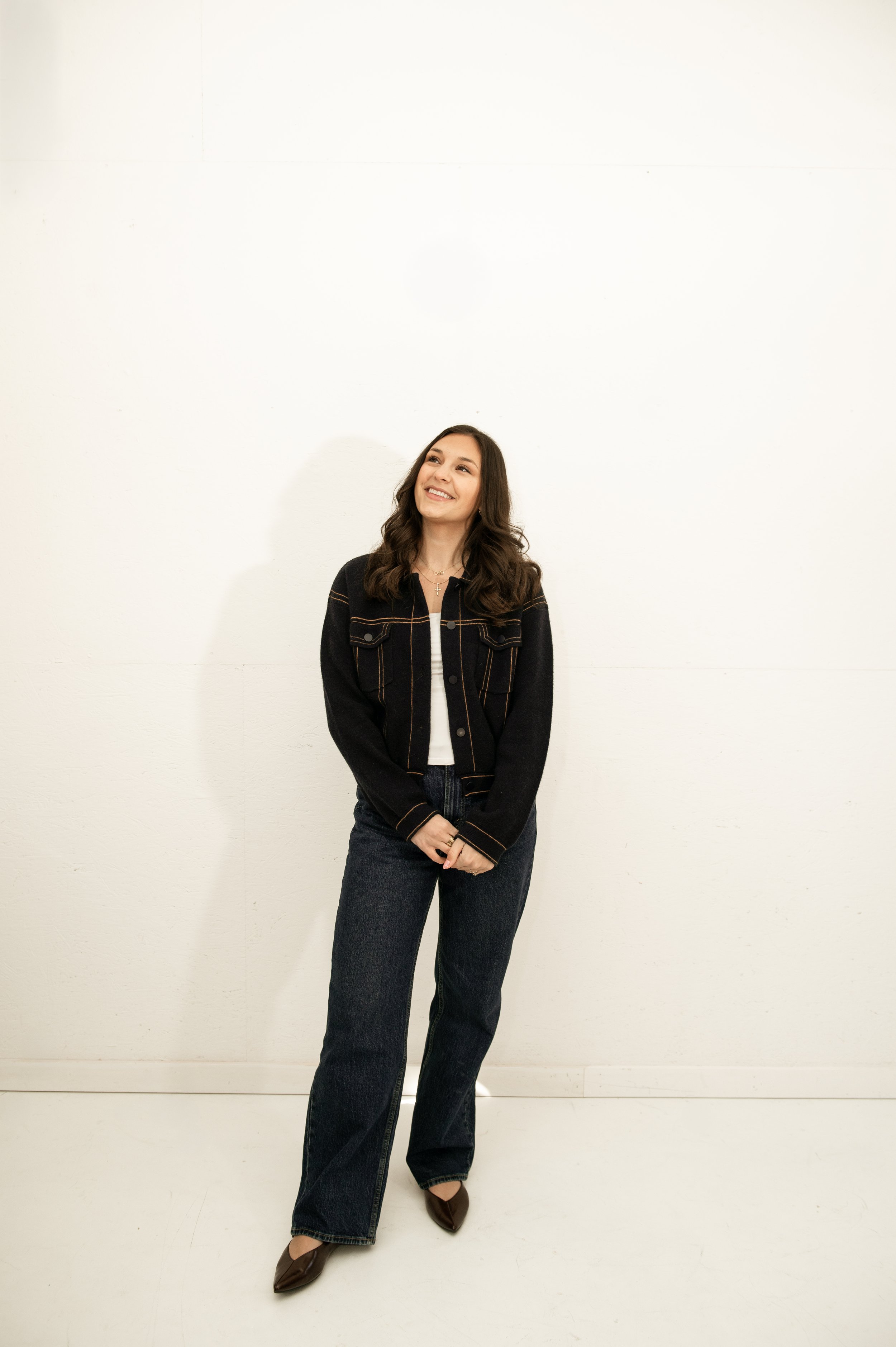 A young woman with long brown hair, wearing a black jacket with brown piping, blue jeans, and brown shoes, smiling and standing against a plain white wall.