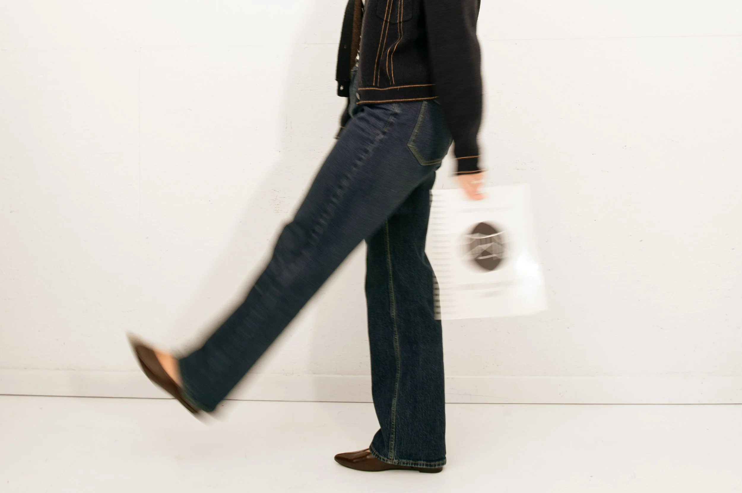 A person walking in a white indoor space, wearing dark jeans, a black jacket, and brown shoes, holding a white shopping bag.
