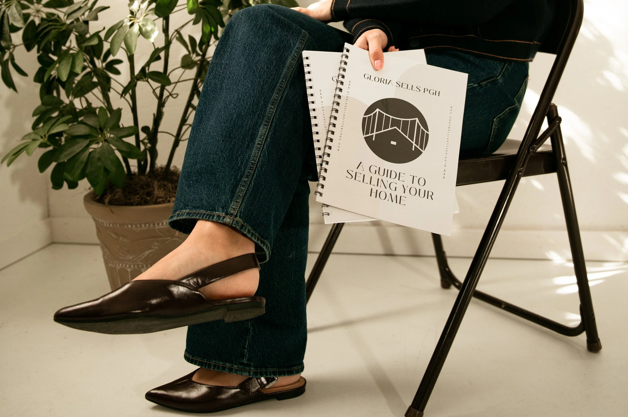 Person sitting on a black chair, holding a guidebook titled "A Guide to Selling Your Home" with a house illustration on the cover. The person is wearing dark jeans, brown shoes, and a black top with orange stitching. Next to the person is a large potted plant with green leaves.