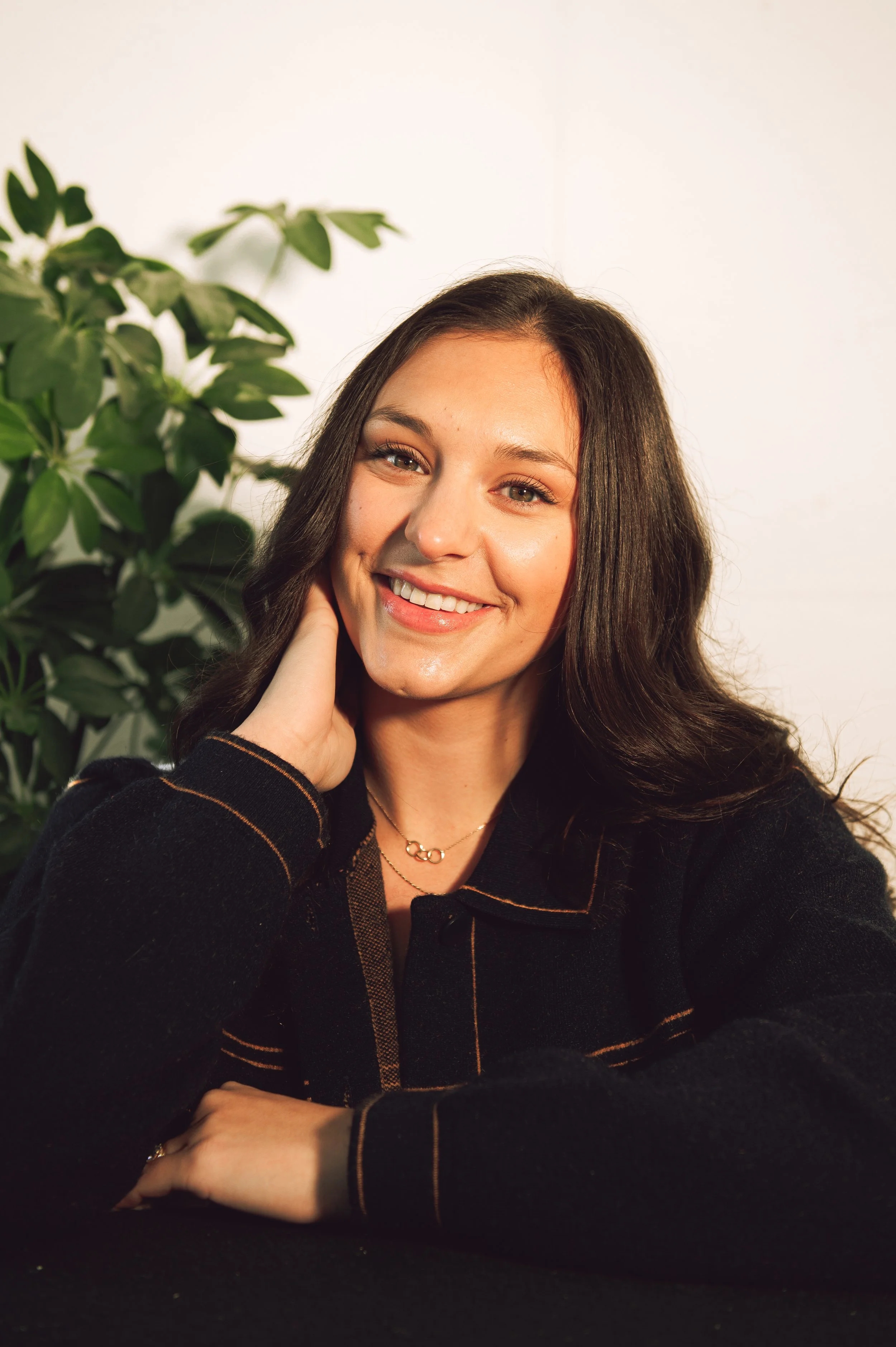 A woman with long brown hair smiling and resting her hand on her neck, sitting at a table with a black surface, with a background of a white wall and a large green plant.