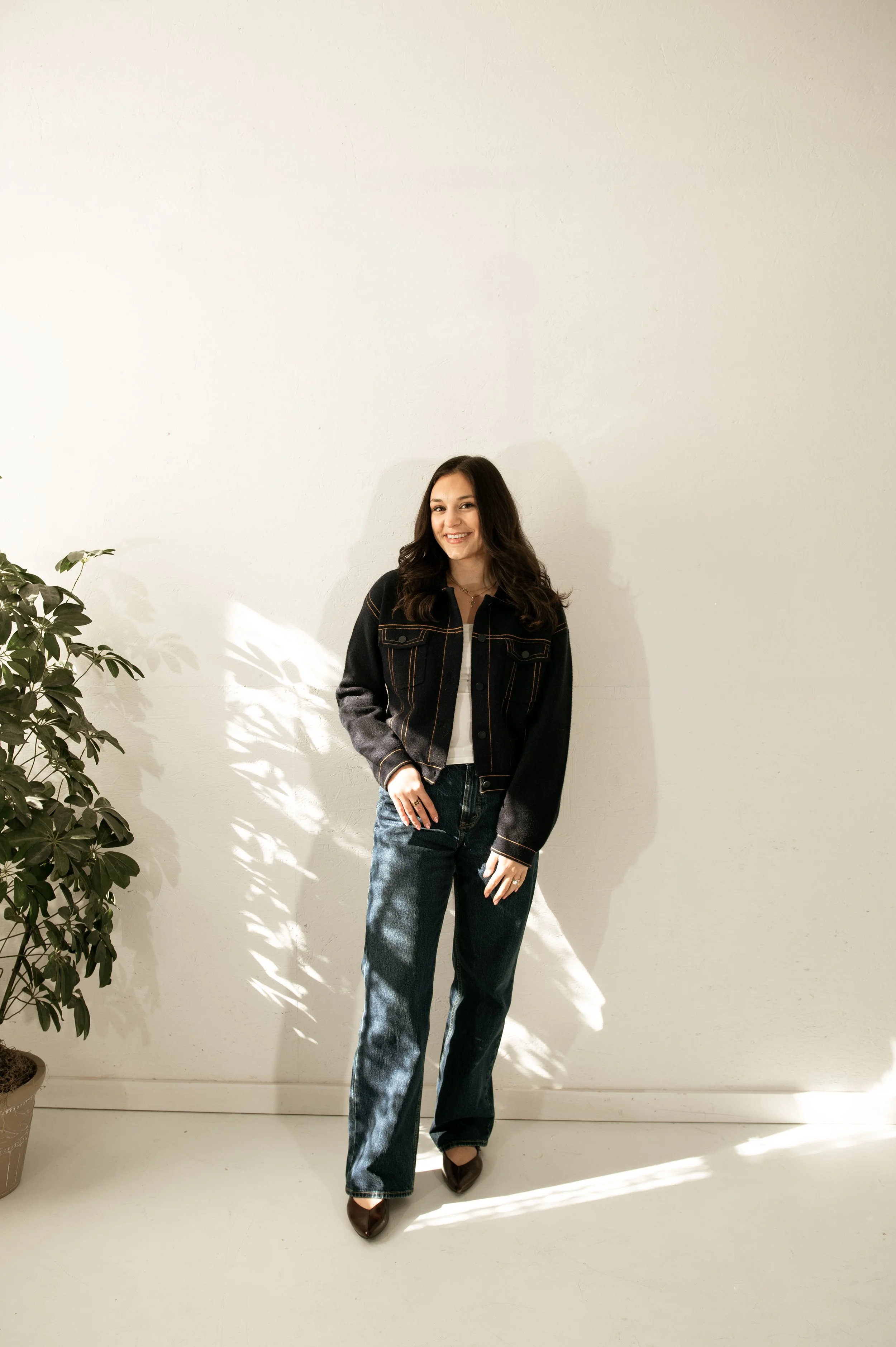 A woman standing against a white wall, smiling, with a potted plant on her left and sunlight casting shadows on the floor.