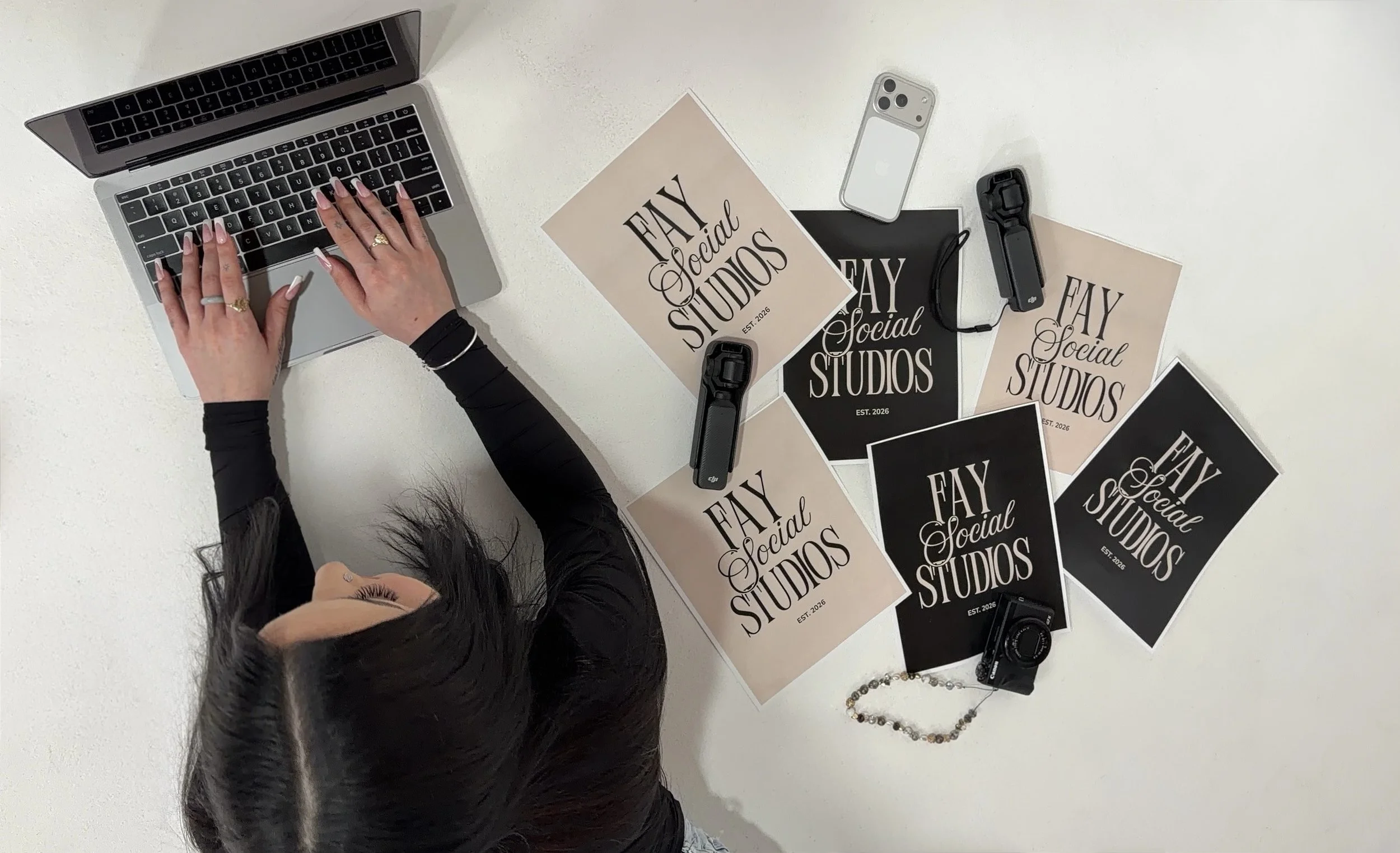 The image shows a woman with long dark hair working at a white table. She is using a laptop with her hands on the keyboard. The table has multiple printed posters with the text 'FAY Social STUDIOS' and 'EST. 2026' in black and beige colors. There are also a smartphone, two cameras, a jewelry necklace, and a beaded bracelet on the table.