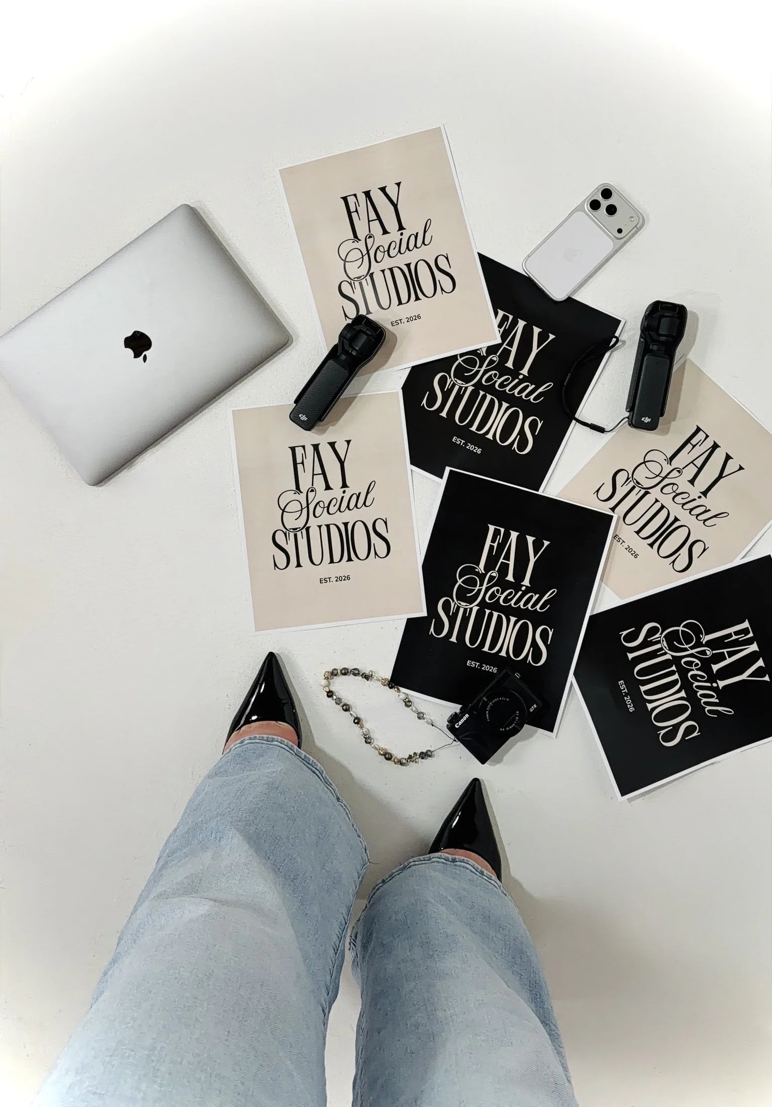 View of a person wearing blue jeans and black shoes photographing a white table from a first-person perspective. The table has multiple Fay Social Studios posters, a closed MacBook, a smartphone, a black wristwatch, and a beaded bracelet.