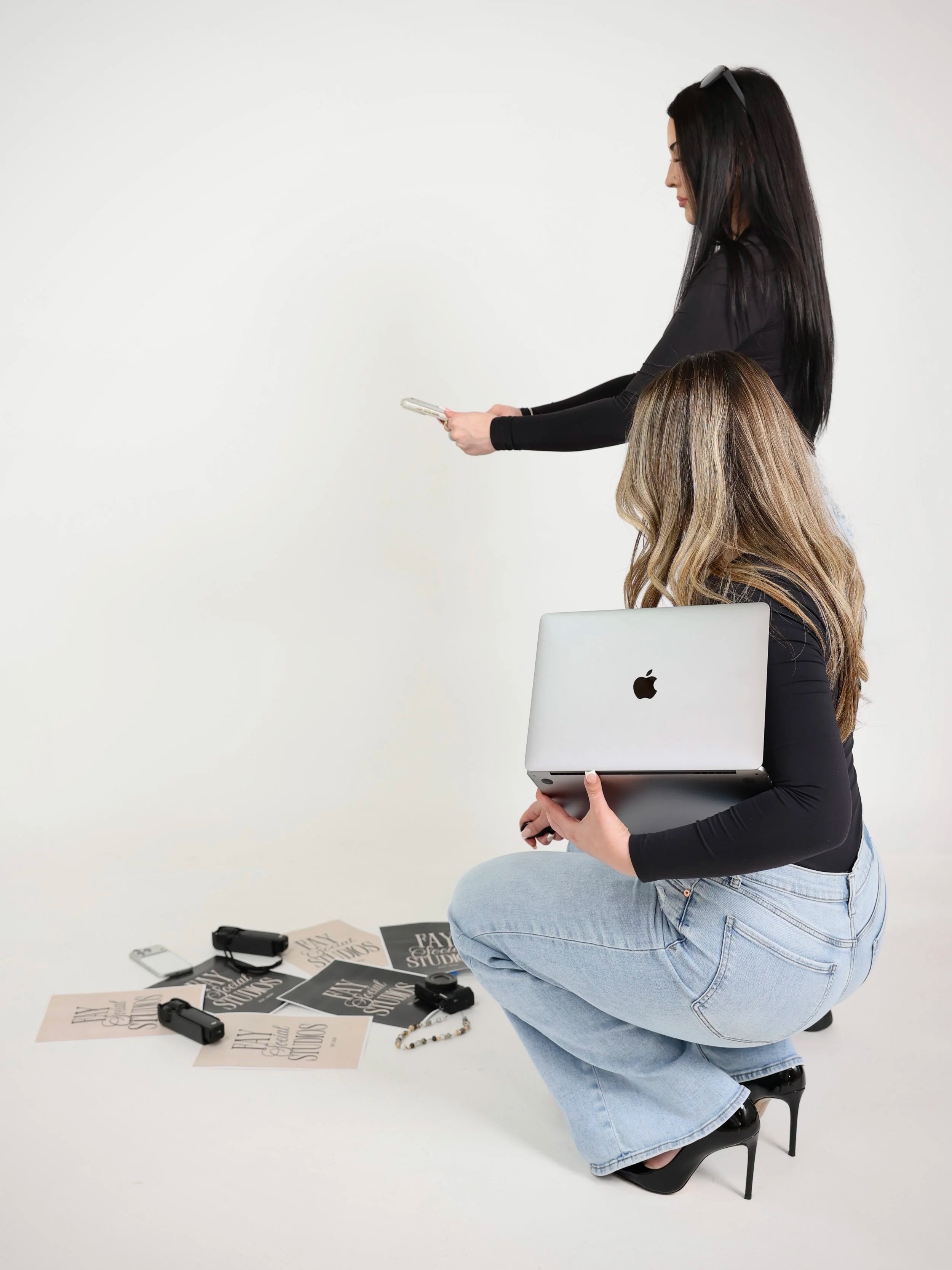 A woman with black hair, wearing glasses on her head, a black long-sleeved top, light blue jeans, and black high heels is kneeling on one knee. She is holding a silver laptop while looking down. There are various printed pages and items on the white floor around her, including a necklace, black clips, and papers with text. Another woman with long brown hair and a black top is standing behind her, holding a smartphone, and looking at the phone.