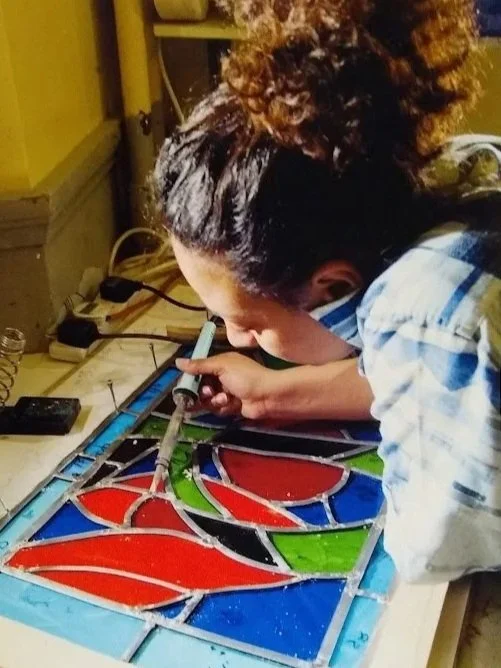 A person with curly hair working on a stained glass art piece on a worktable, holding a soldering iron.
