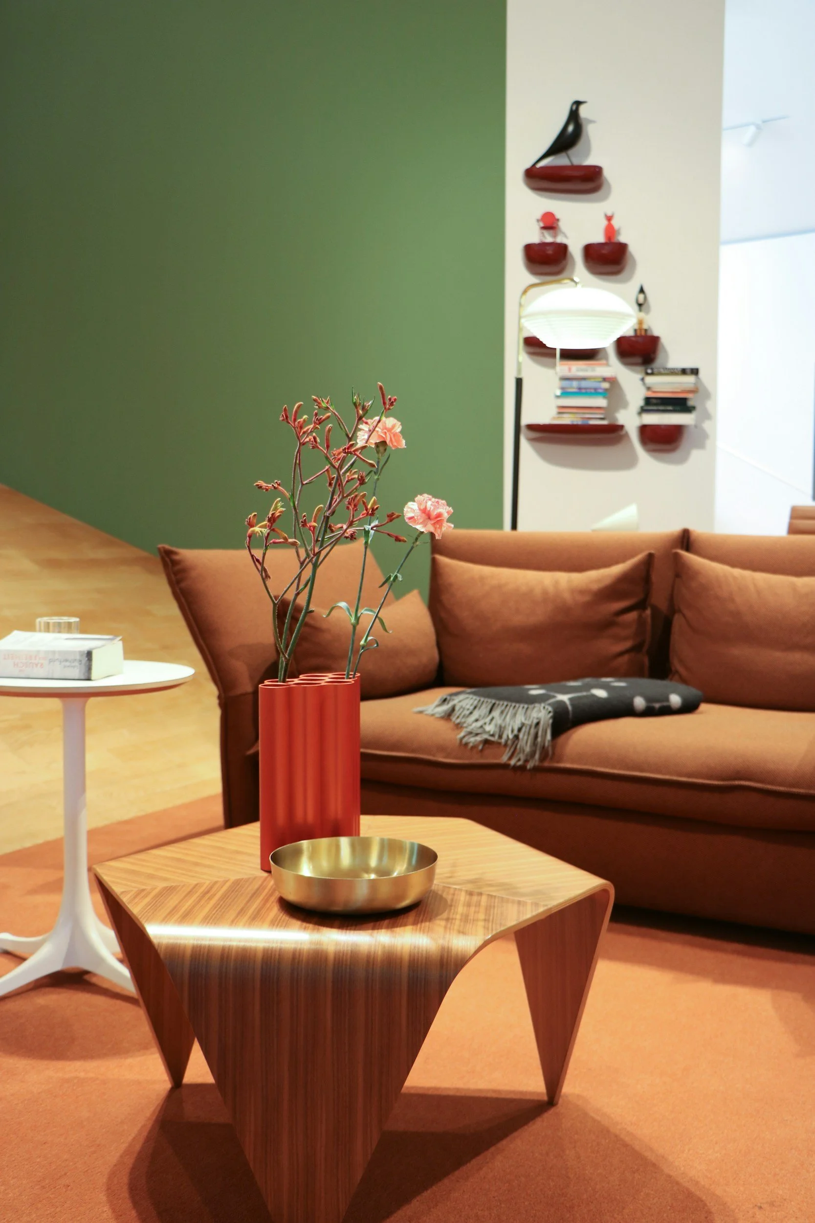 A living room with a brown sofa, a wooden coffee table with a gold bowl and a tall orange vase with pink flowers. There is a green and white wall with floating red shelves holding books, decorative objects, and a lamp. Part of a white lamp with a curved stand is also visible.