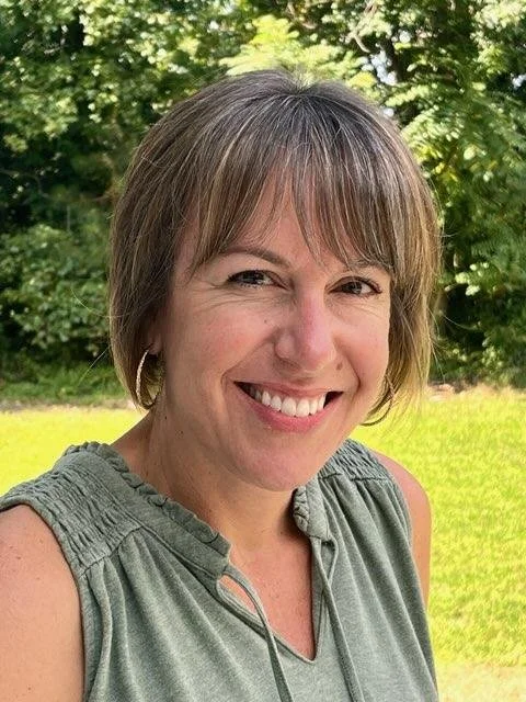 A woman with short brown hair smiling outdoors with green trees and grass in the background.