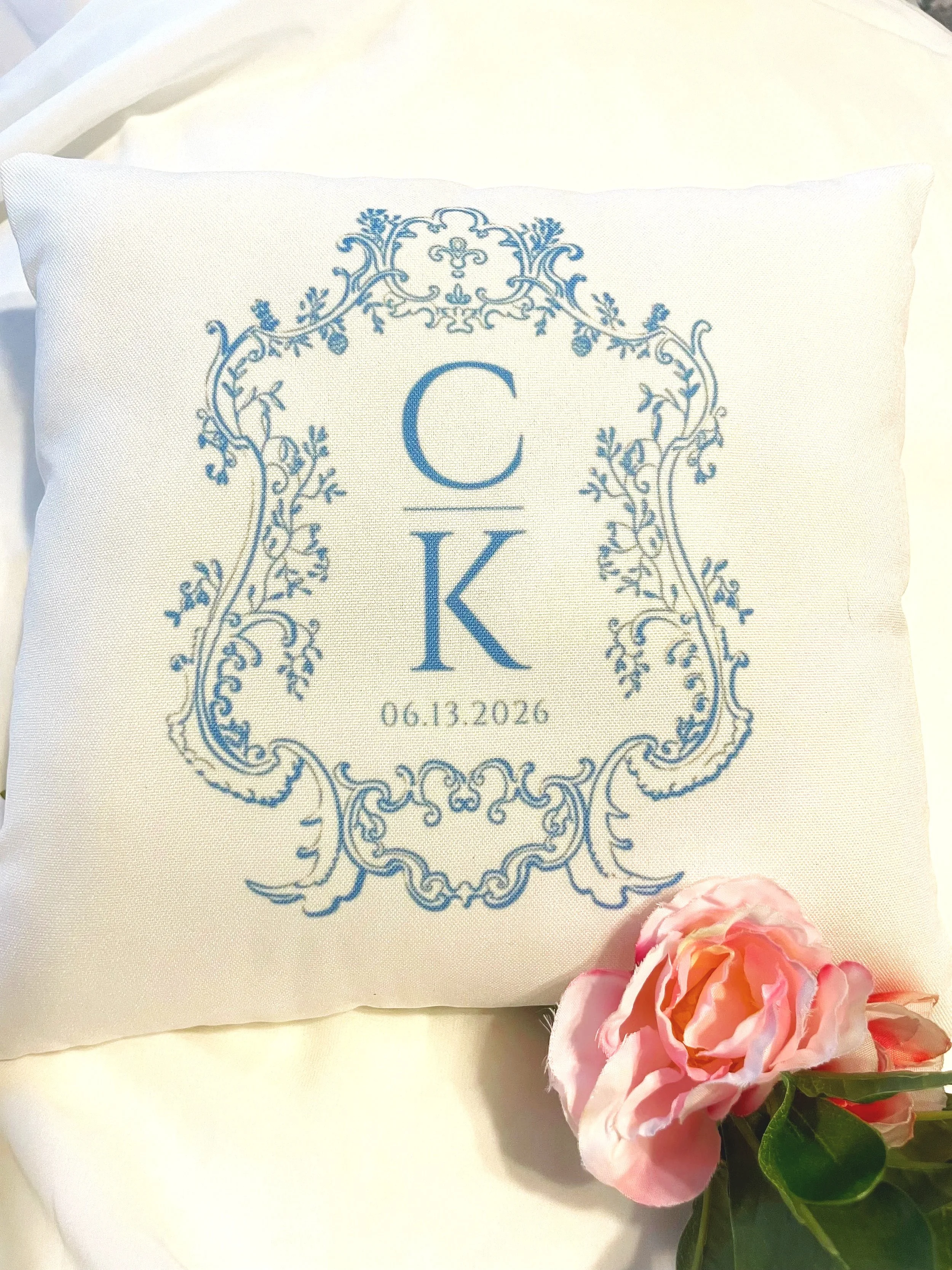 A white pillow with blue embroidered monogrammed initials 'C K' and a date '06.13.2026' inside an ornate blue frame, with pink artificial flowers placed beside it.