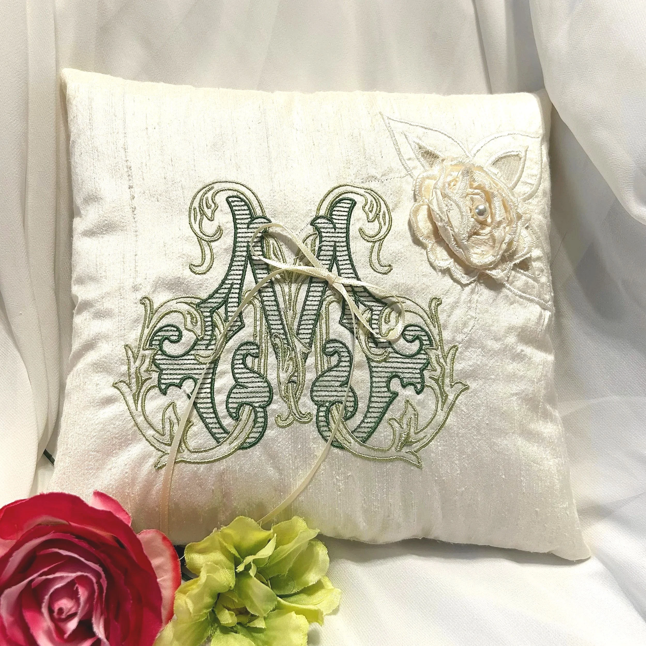 A white embroidered pillow with a floral design featuring roses, roses and ribbon details, placed in front of artificial red, yellow, and green flowers on a light-colored fabric background.