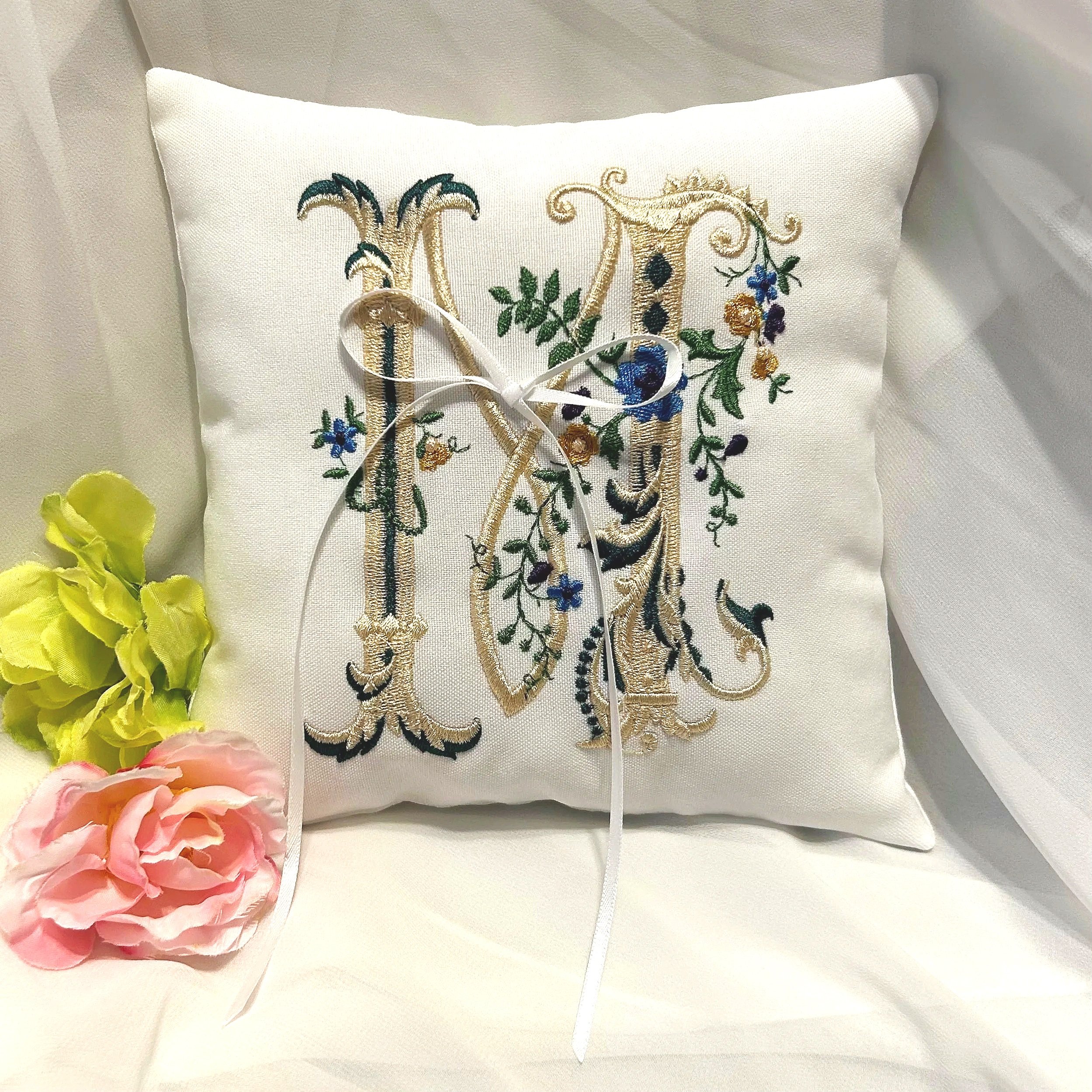 Embroidered pillow with floral and flourish design in gold, green, and blue, with yellow and pink flowers nearby.