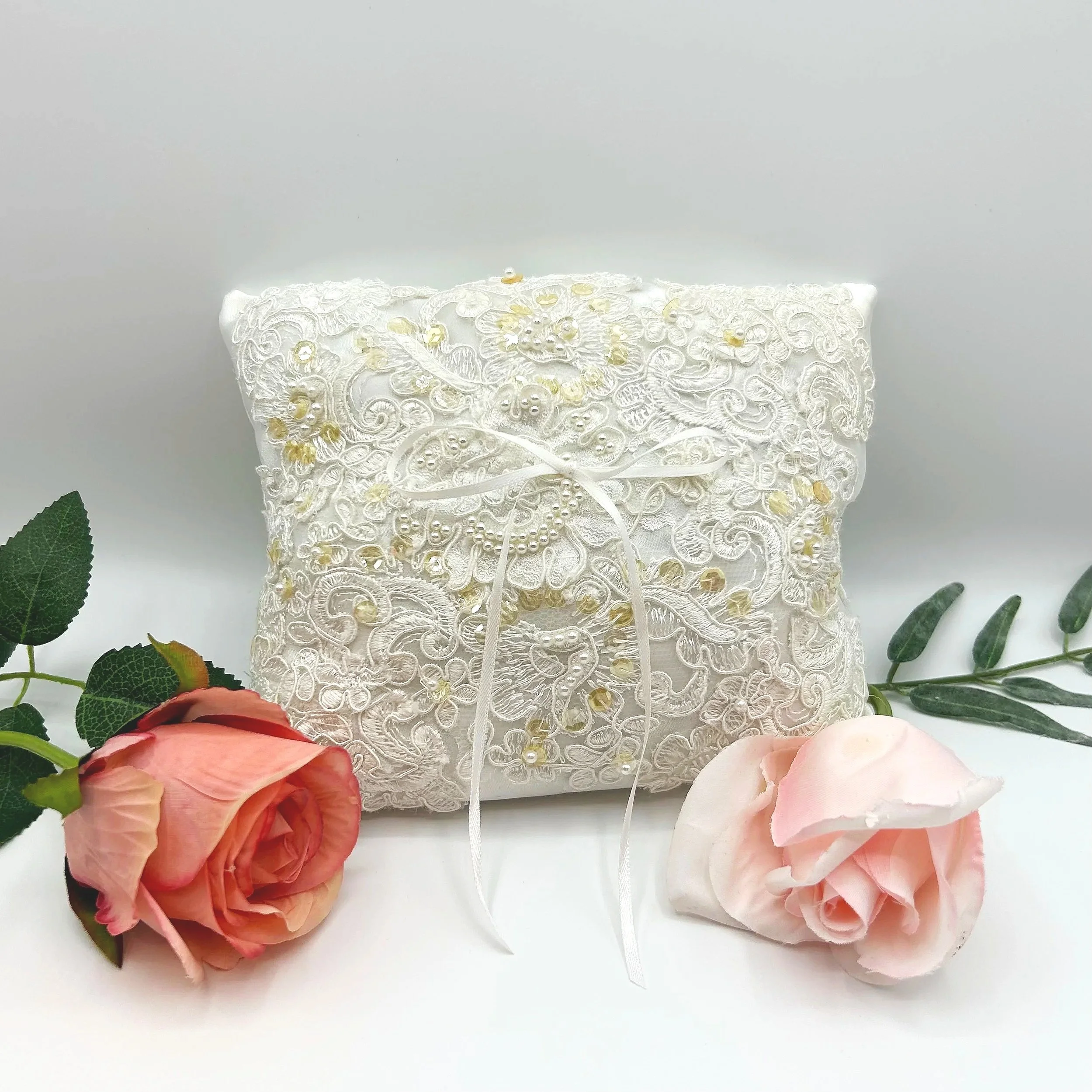 Intricate white lace fabric with gold embroidery and small pearl accents, surrounded by pink and peach roses with green leaves.