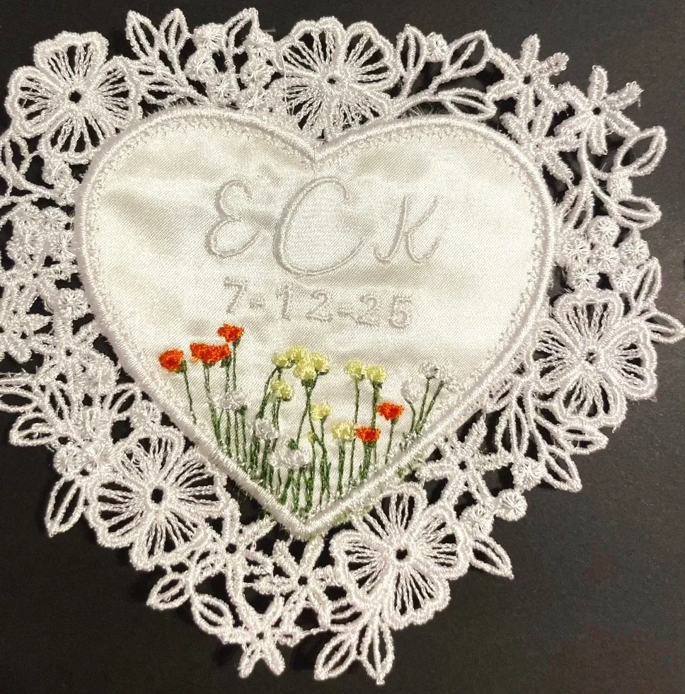 A heart-shaped embroidered cloth with a floral lace border. The embroidery features initials 'ECK', a date '7-12-25', and an embroidered garden of orange and yellow flowers at the bottom.