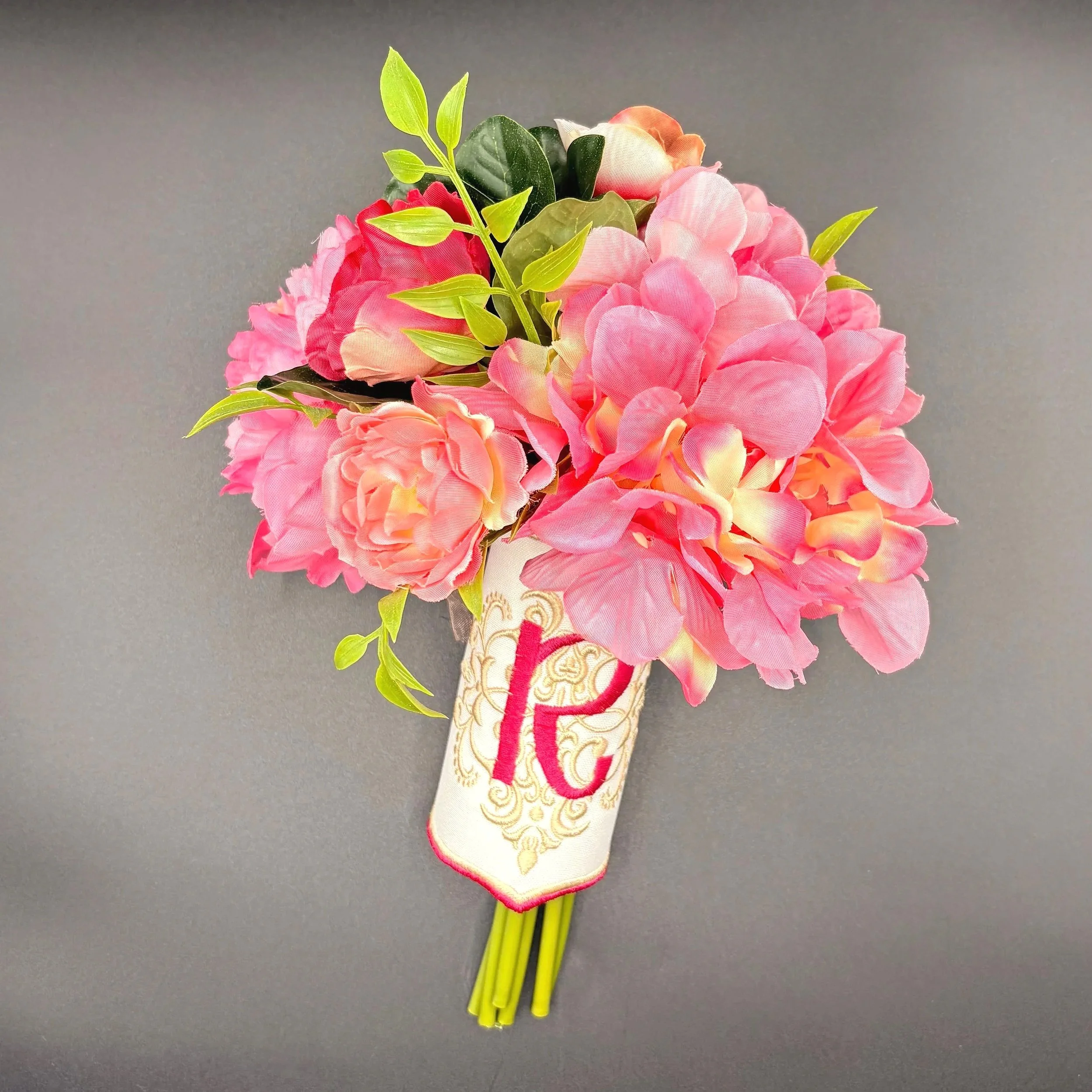 A small artificial bouquet of pink hydrangea flowers with green leaves, wrapped in decorative fabric with a letter 'G' on it, set against a dark gray background.