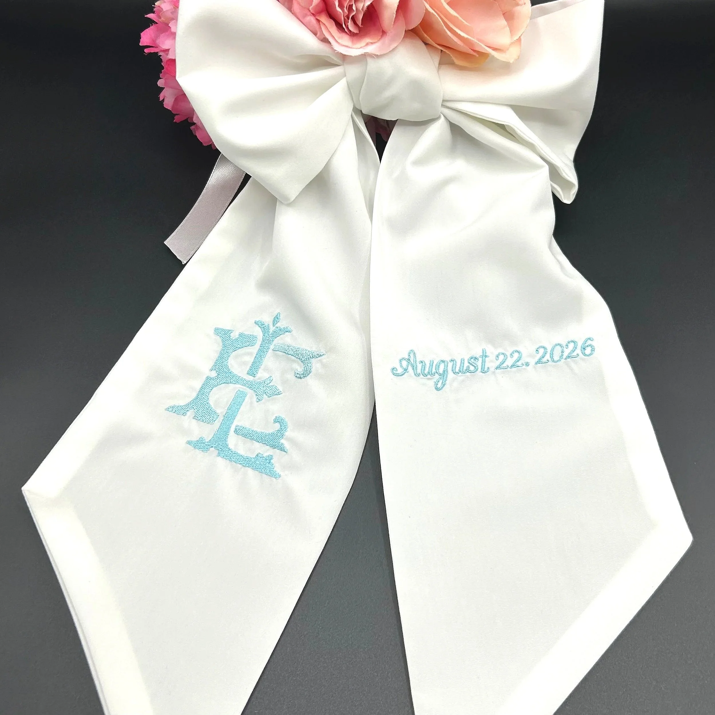A close-up of a white cloth with embroidered blue tree and date, with pink flowers and a white bow on top, on a dark surface.