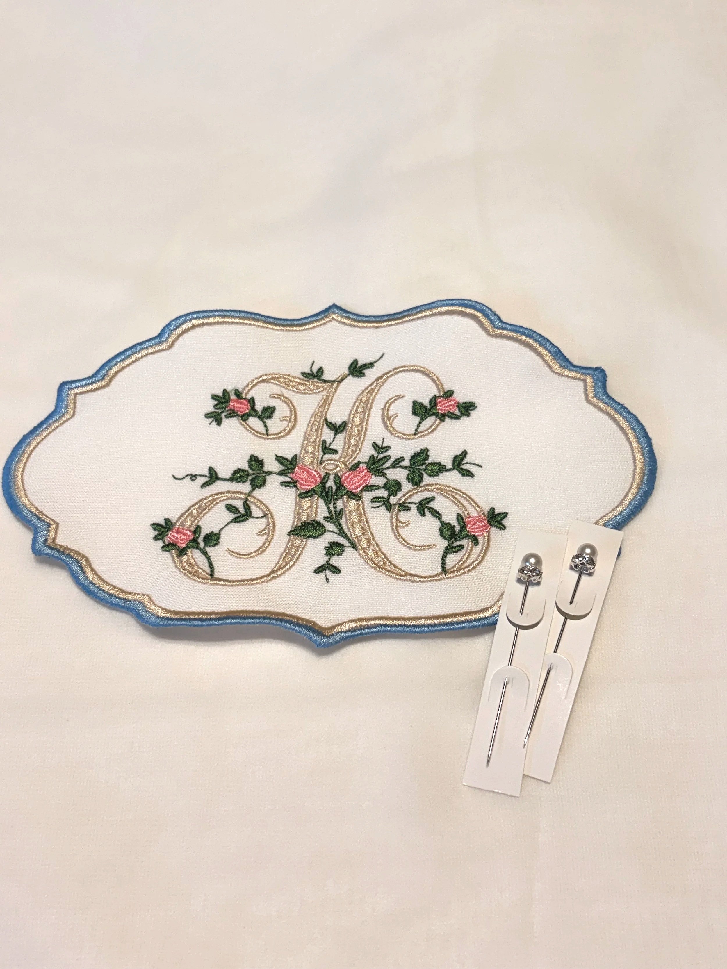 Embroidery piece with floral design and monogram, two sewing pins on white backing.