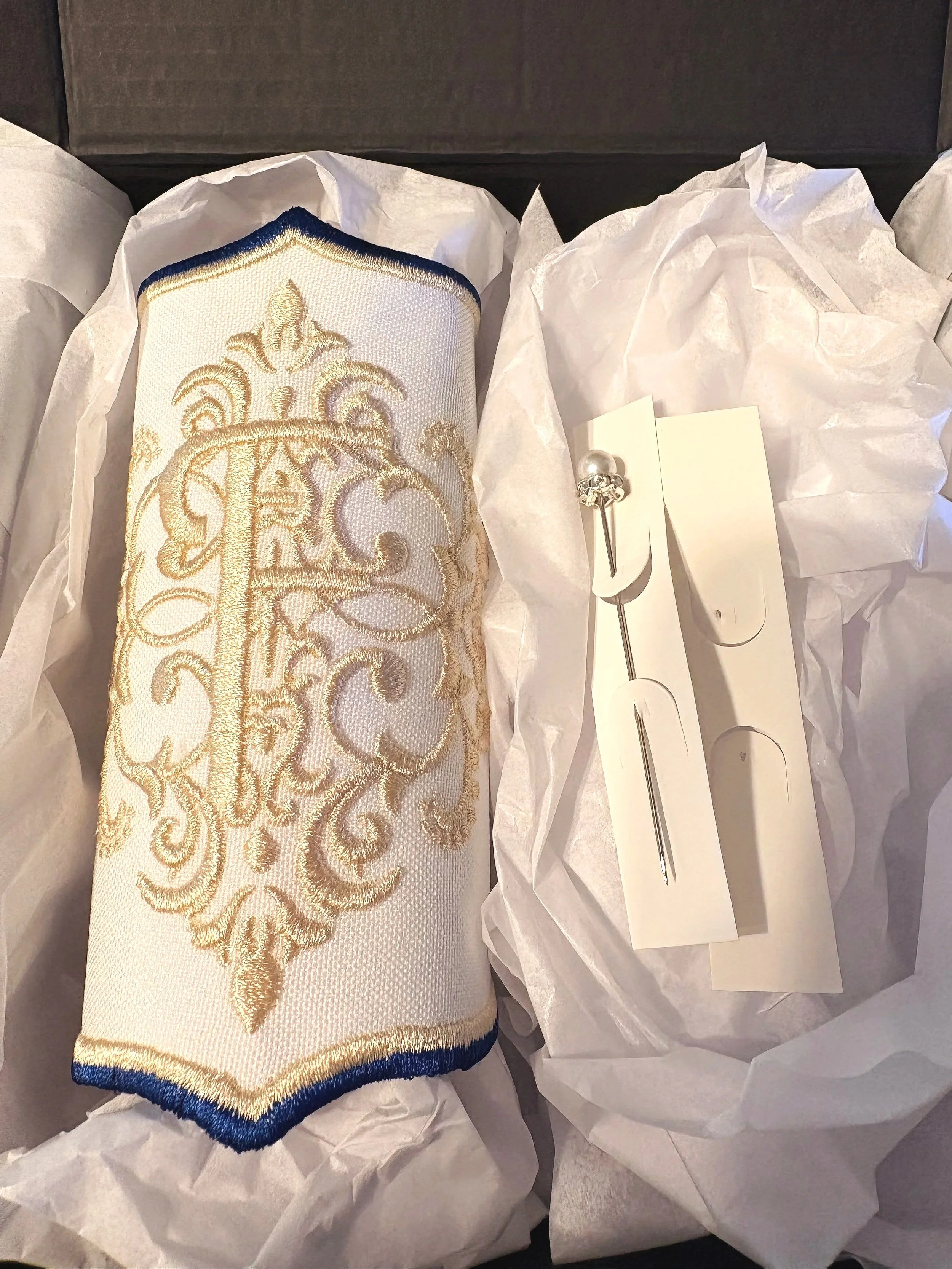 A gold and white embroidered piece, possibly a decorative pillow or fabric, surrounded by white tissue paper. There is also a small silver pin and a white box nearby.