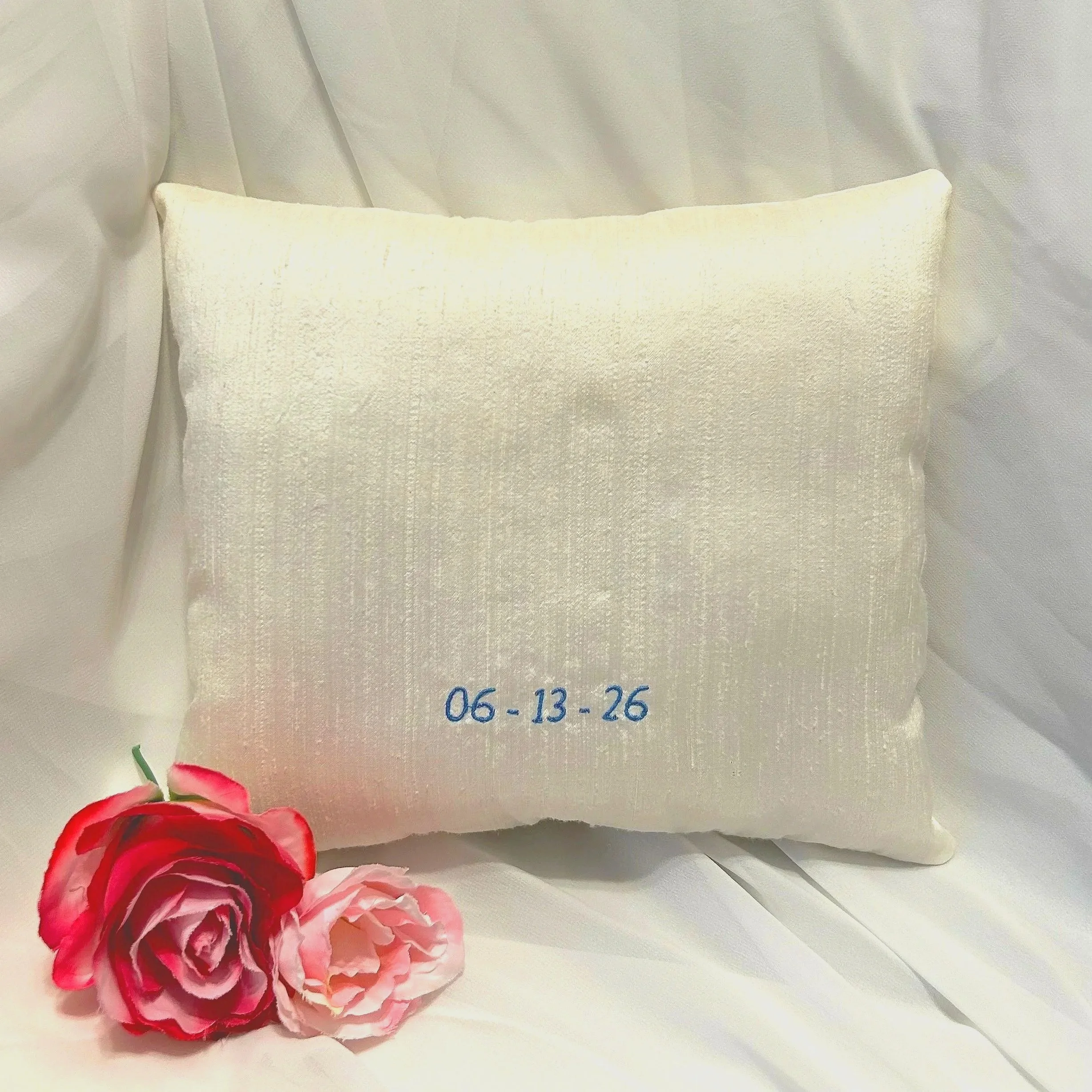 A close-up of two pink and white flowers, possibly carnations, positioned on a white textured background with some fabric. The date 06-13-26 is embroidered in blue on the background.