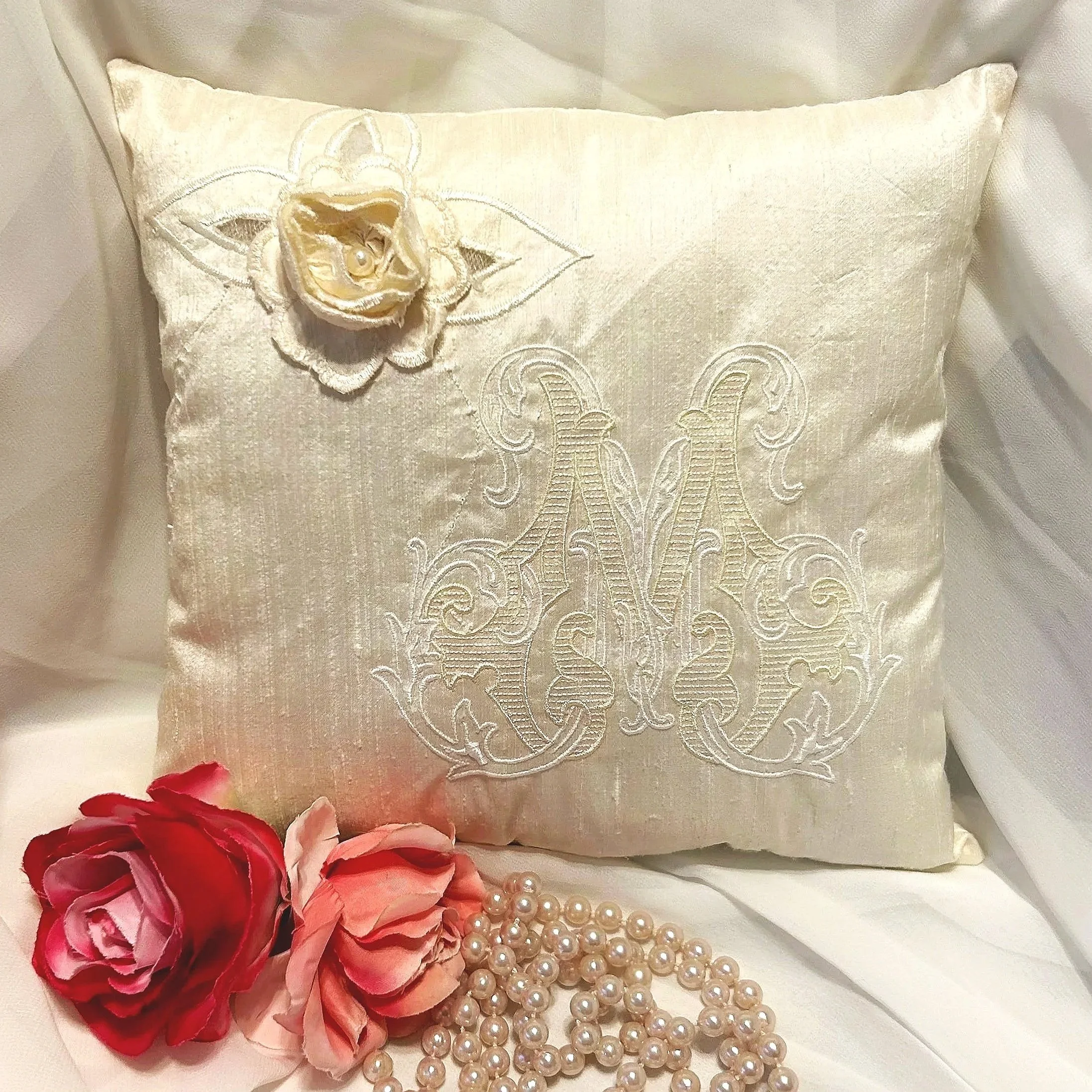 A decorative pillow with cream-colored fabric, embroidered with a butterfly and a rose design, giving it an elegant appearance. Surrounding the pillow are pink and red artificial flowers and a strand of pearl beads, creating a soft and luxurious sett