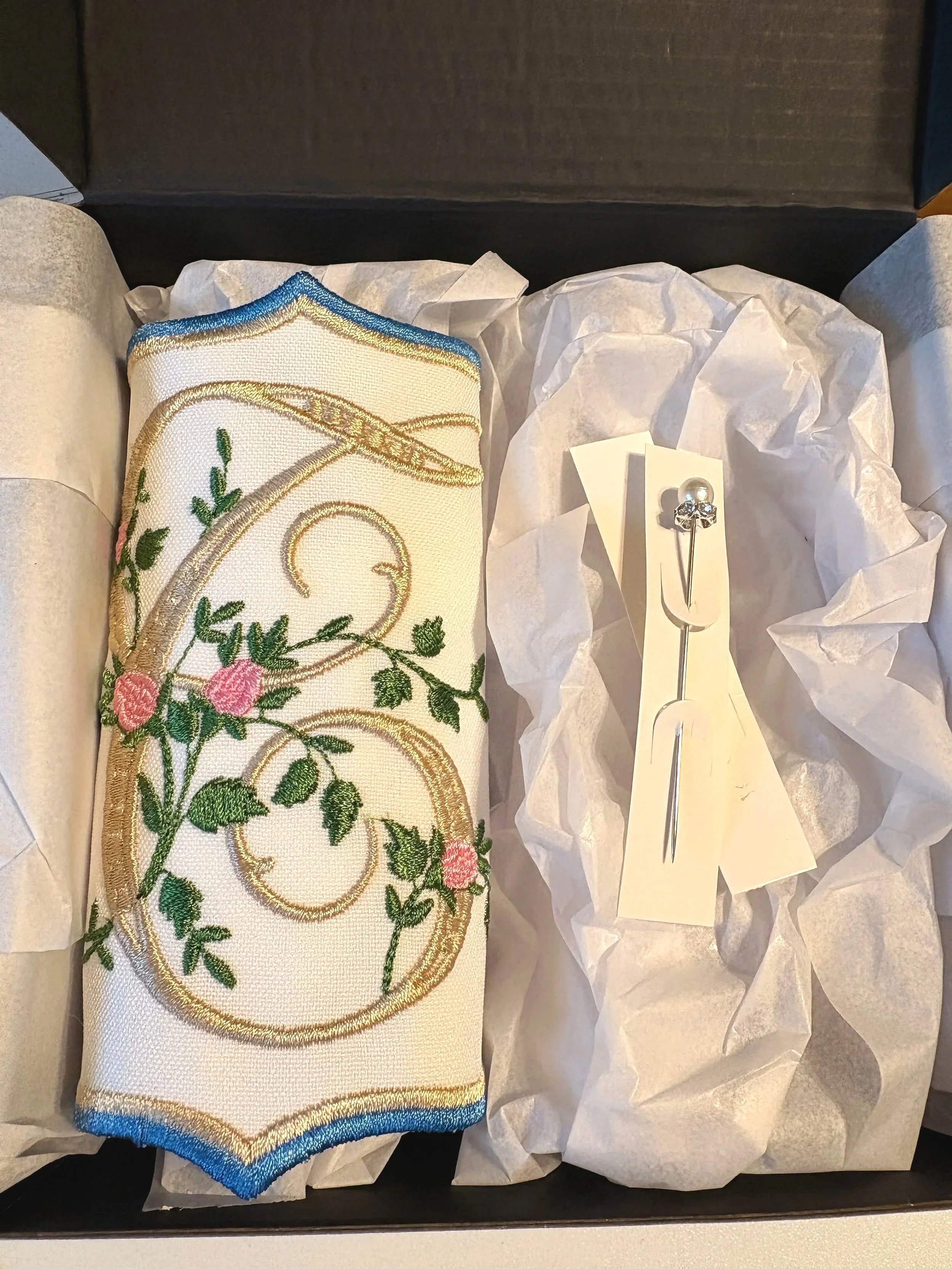 A decorative embroidered patch with floral design and gold accents, inside a black box with tissue paper and a pin with a skull design.