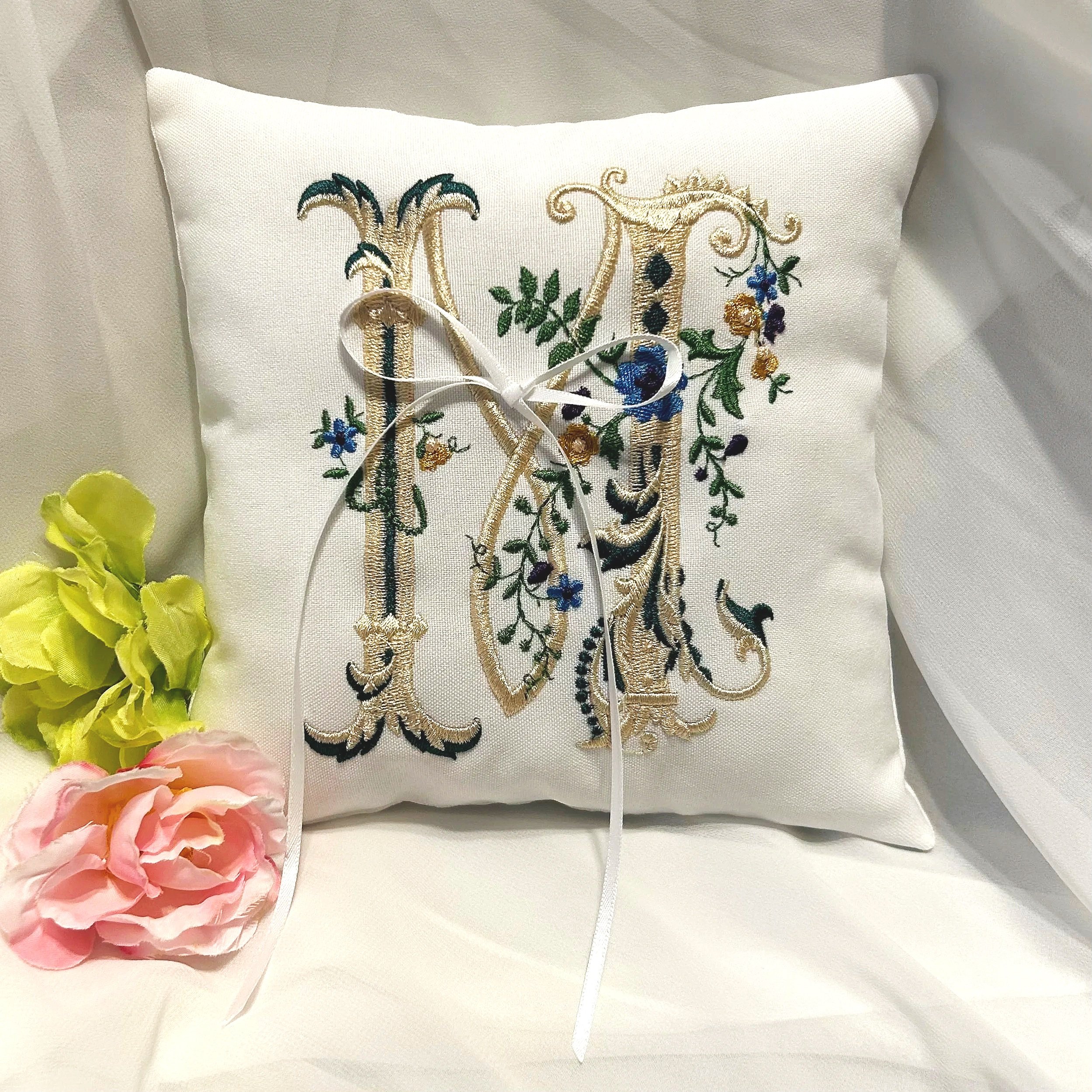 A white embroidered pillow with gold, black, green, and blue intricate floral and design details, tied with a white ribbon, and decorated with pink and yellow flowers on the side.