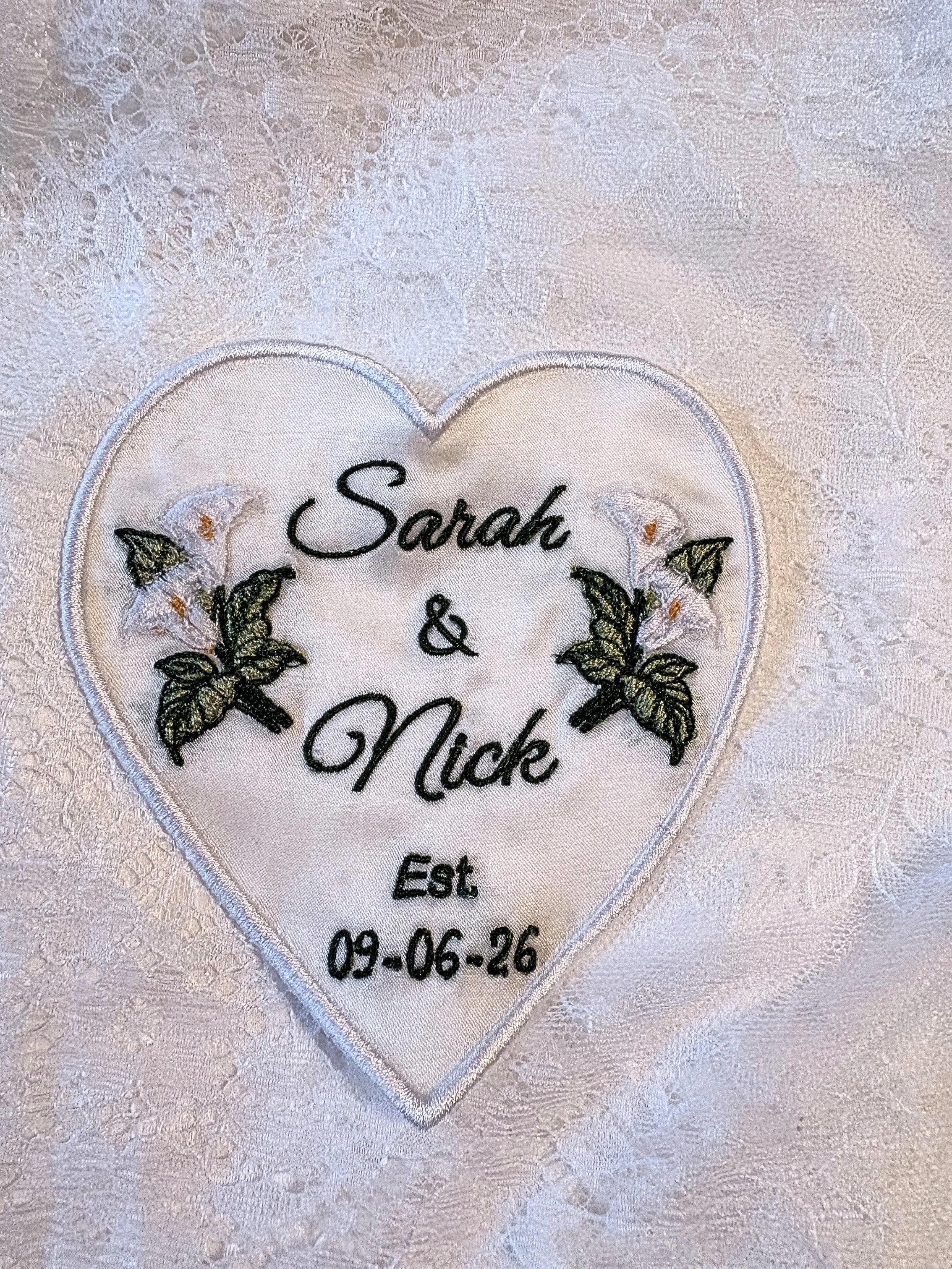 Close-up of embroidered white heart-shaped fabric with names Sarah and Nick, wedding date 09-06-26, on textured background.