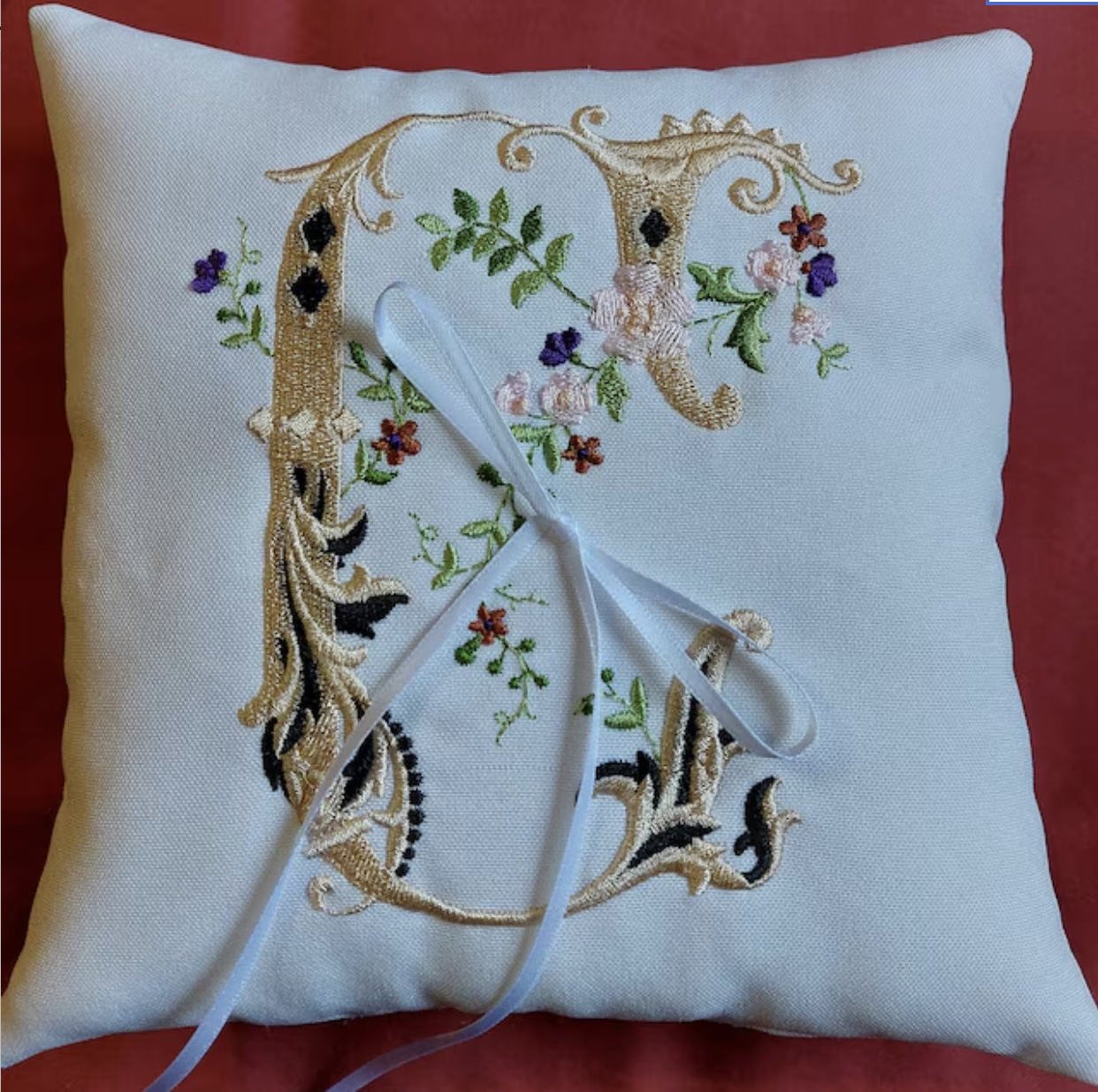 A white embroidered pillow with a floral letter 'G' and a white ribbon tied in a bow.