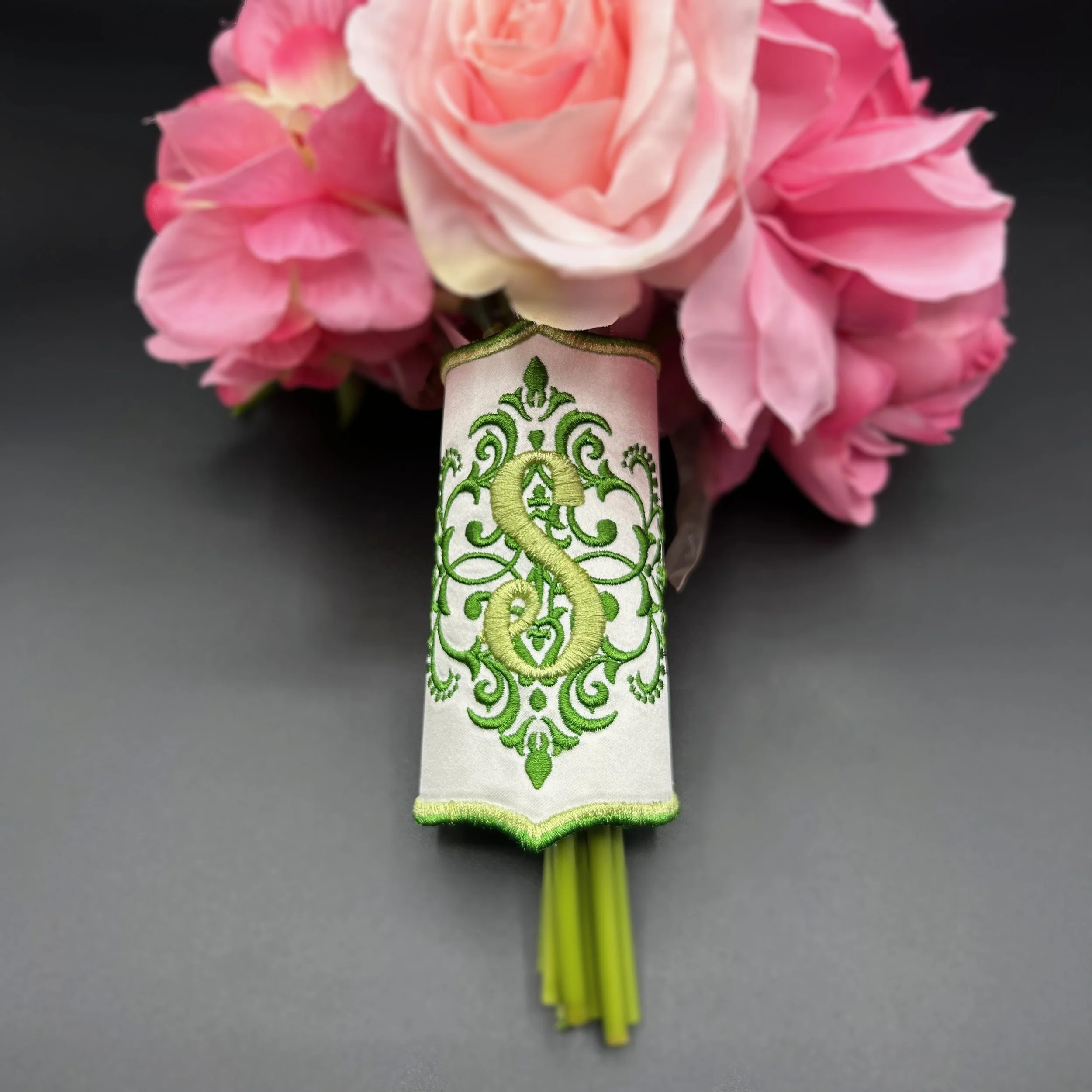 Close-up of a pink and white flower boutonniere with embroidered green and gold design on the ribbon at the base, against a black background.