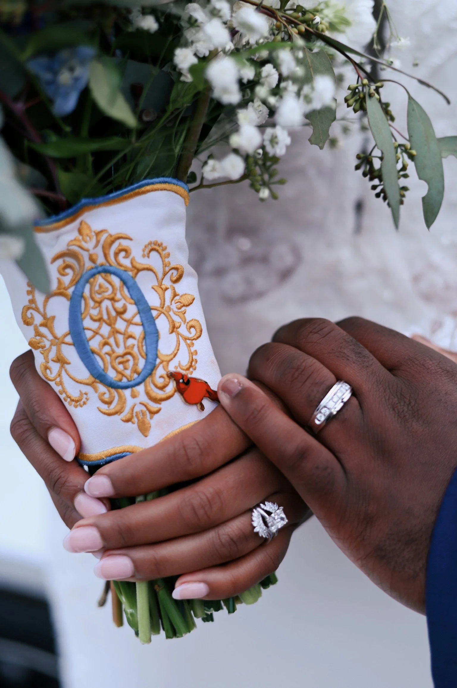 Two hands are holding a bouquet of white flowers with green leaves, wrapped in a cloth with embroidered design.