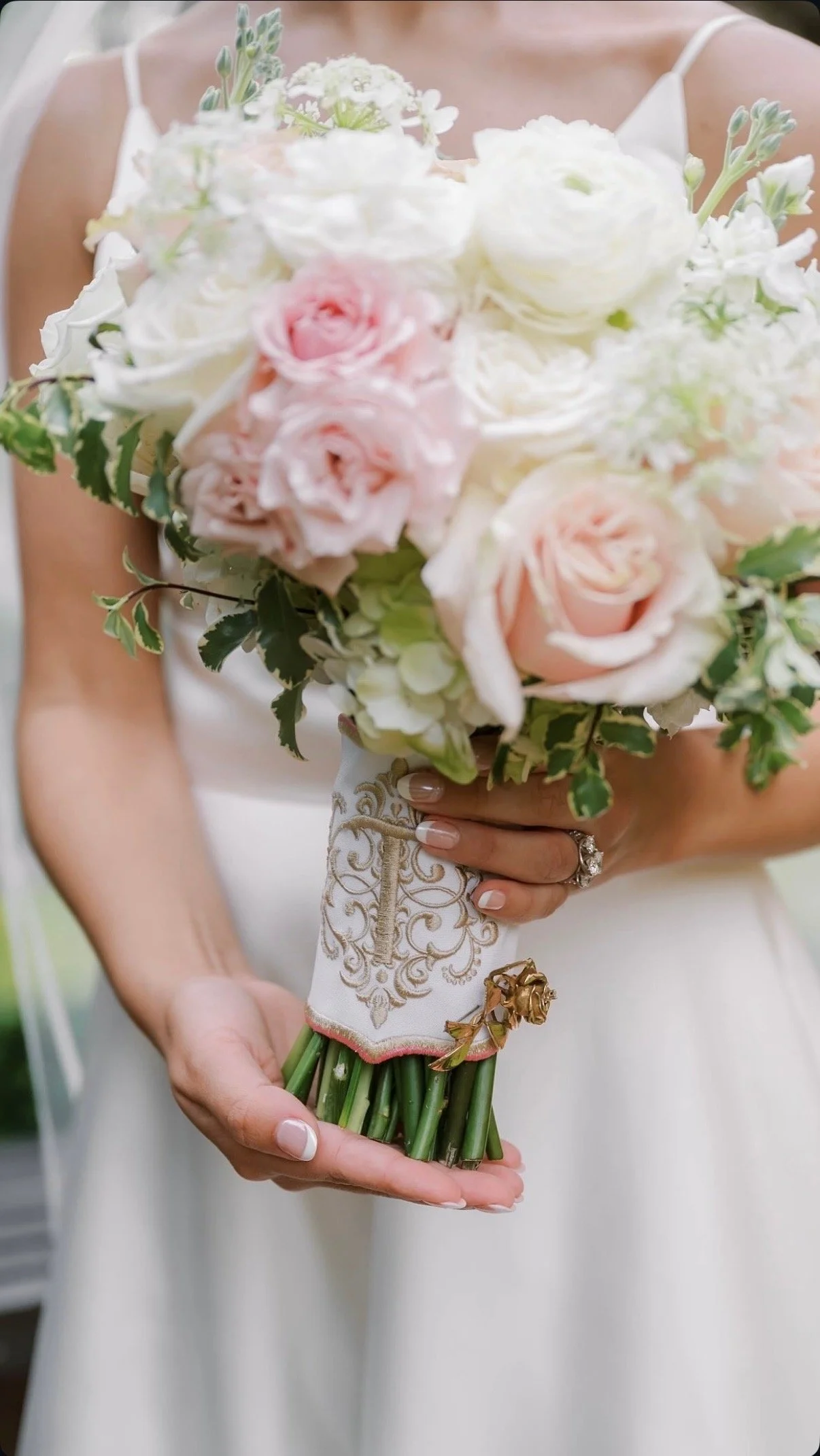 A bride in a white dress holds a bouquet of pink and white roses with green leaves.