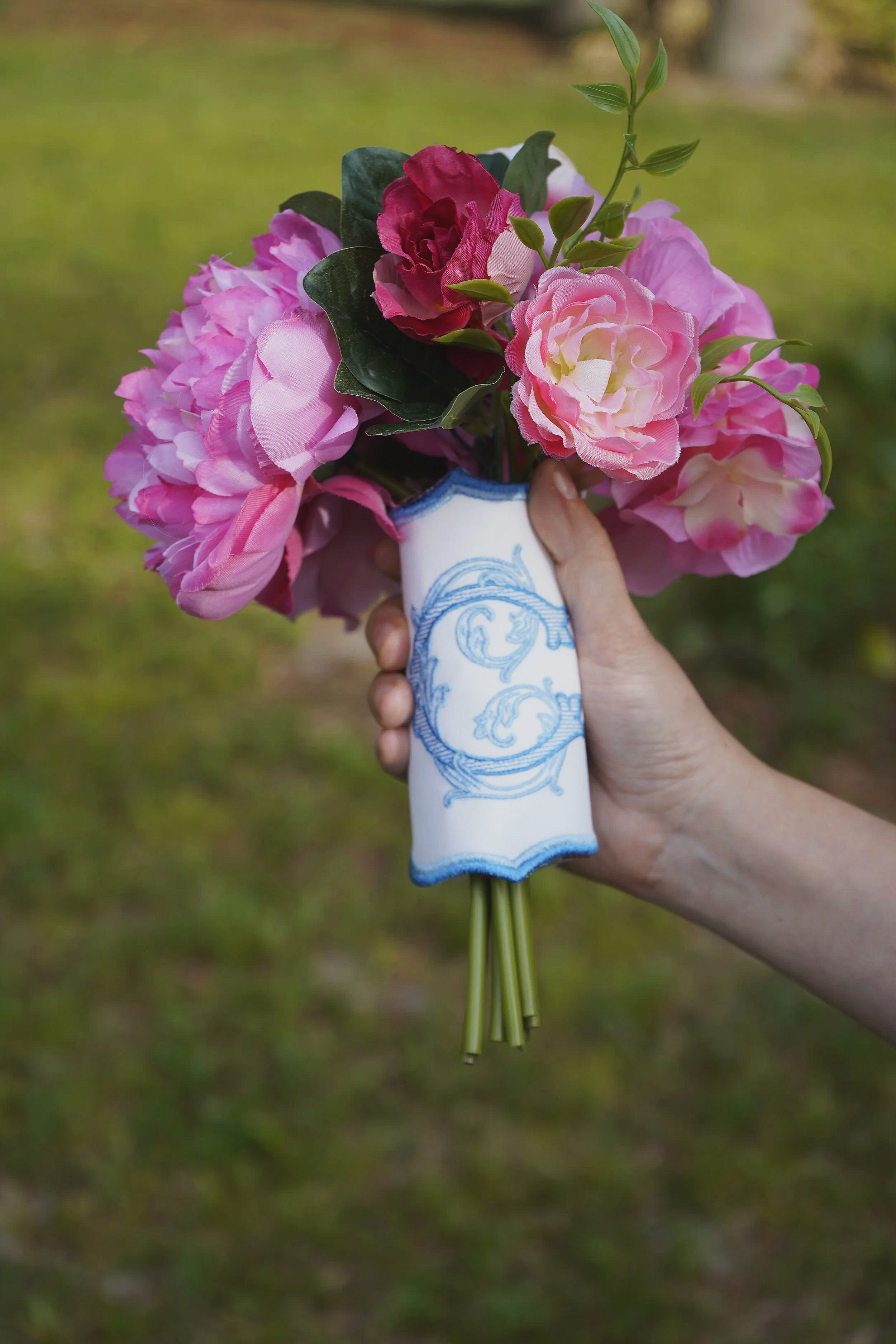 Person holding a bouquet of pink and red artificial flowers with green leaves in a vase with blue decorative patterns.