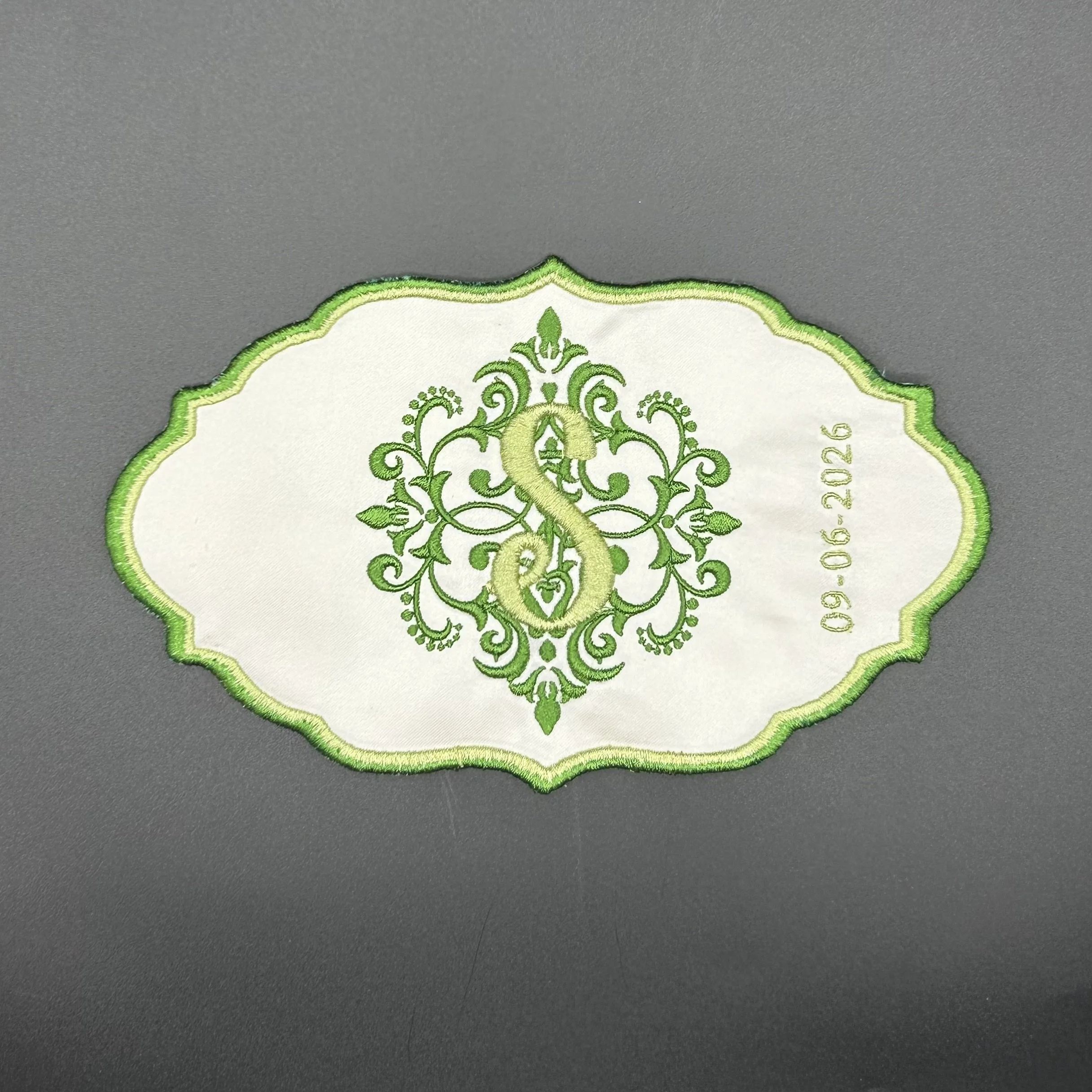 Close-up of embroidered patch with intricate green and yellow design on white fabric, bordered with green stitching, resting on a gray surface.