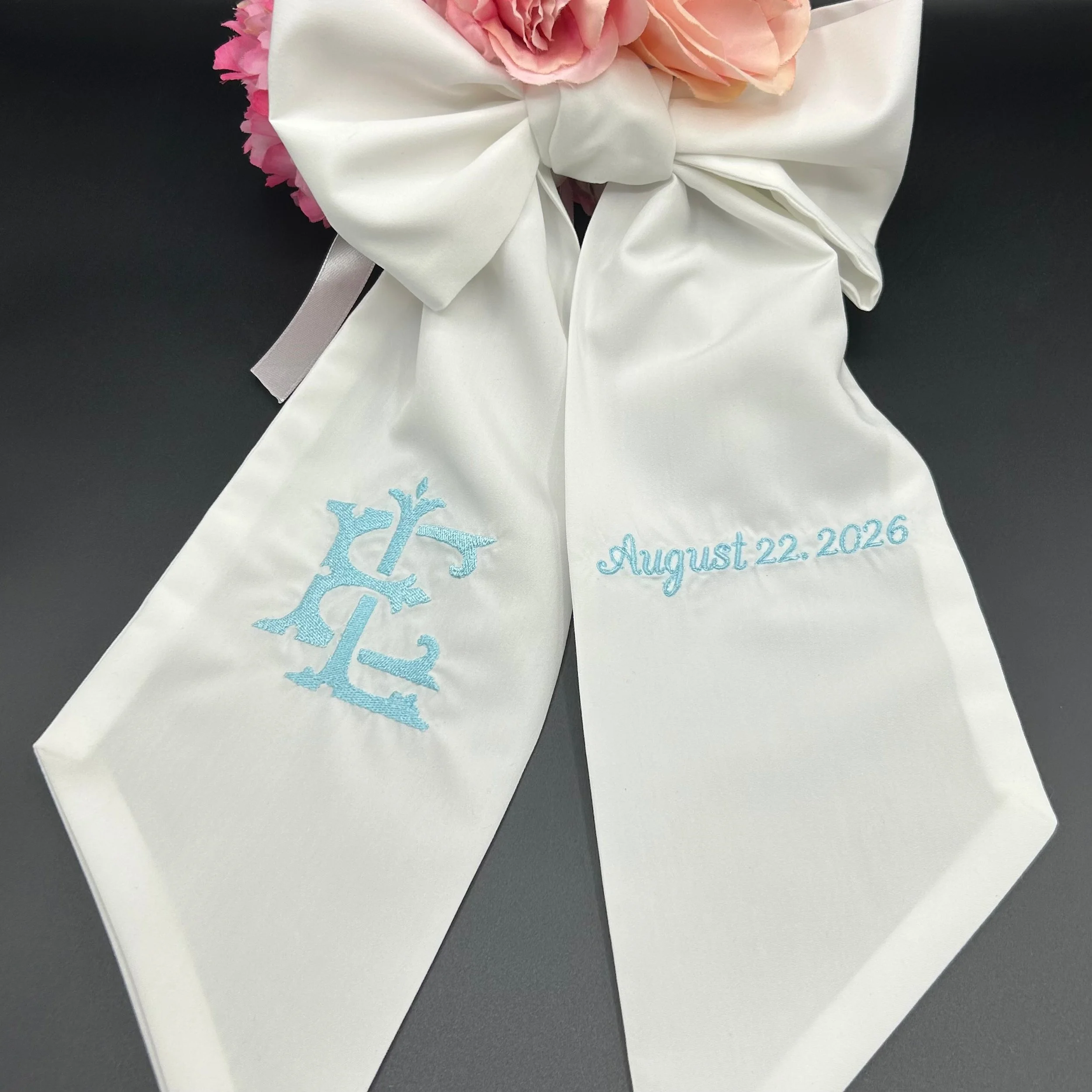 A white satin bag with embroidered blue tree design and the date August 22, 2026, visible on the front. The bag is tied with a white satin ribbon and decorated with pink and peach roses. The background is black.