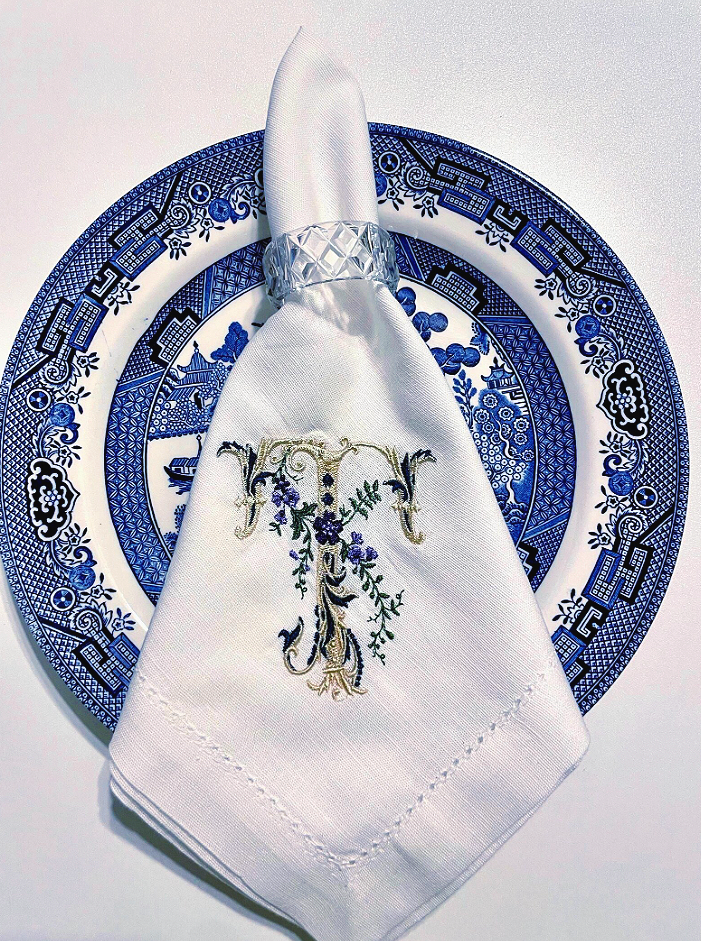 A white cloth napkin with embroidered details, placed on a blue and white decorative plate, with a clear glass napkin ring.