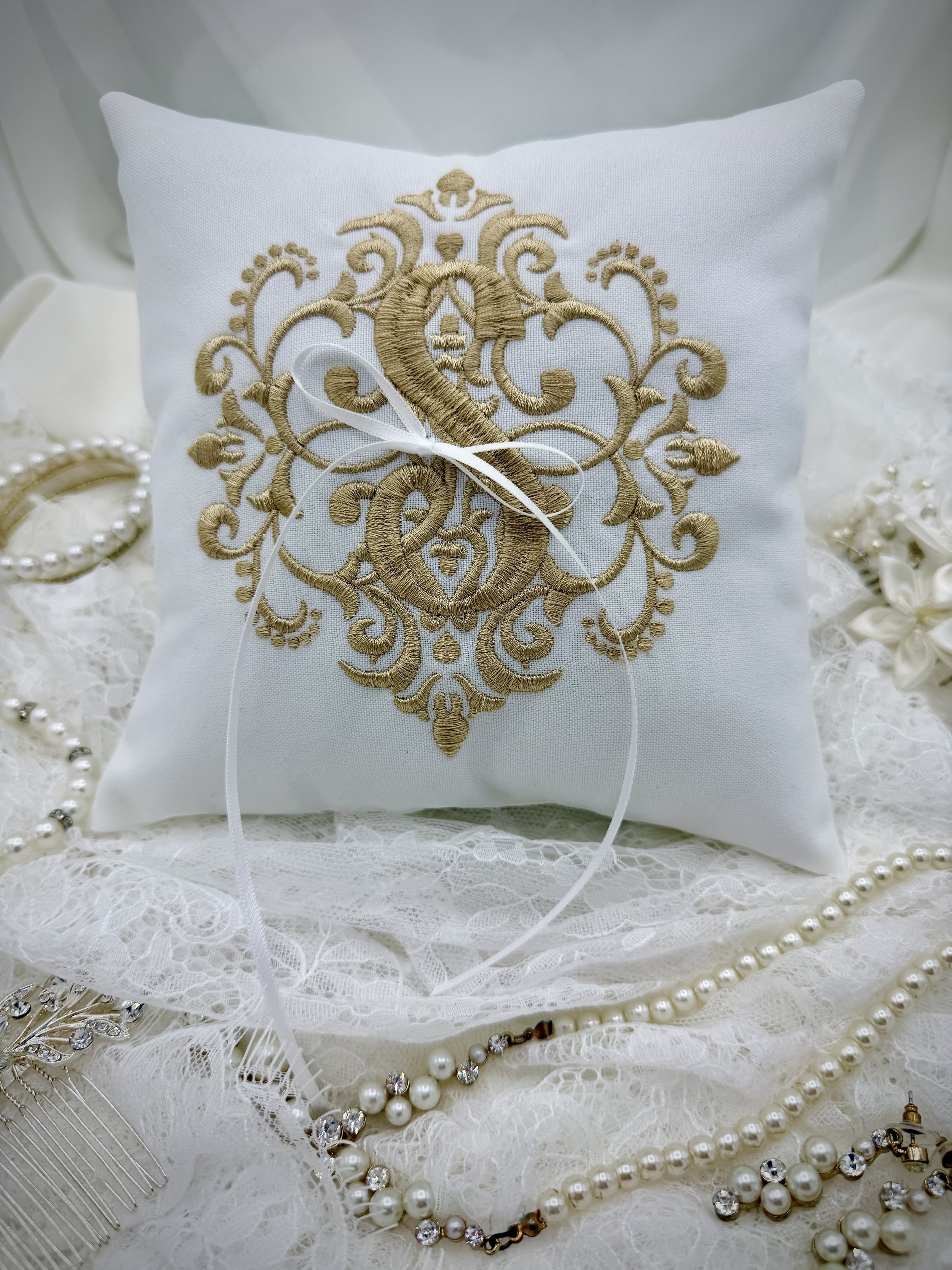 A white pillow with gold embroidery featuring an ornate crest design, surrounded by jewelry including pearl necklaces, earrings with rhinestones, a hair comb, and lace fabric.