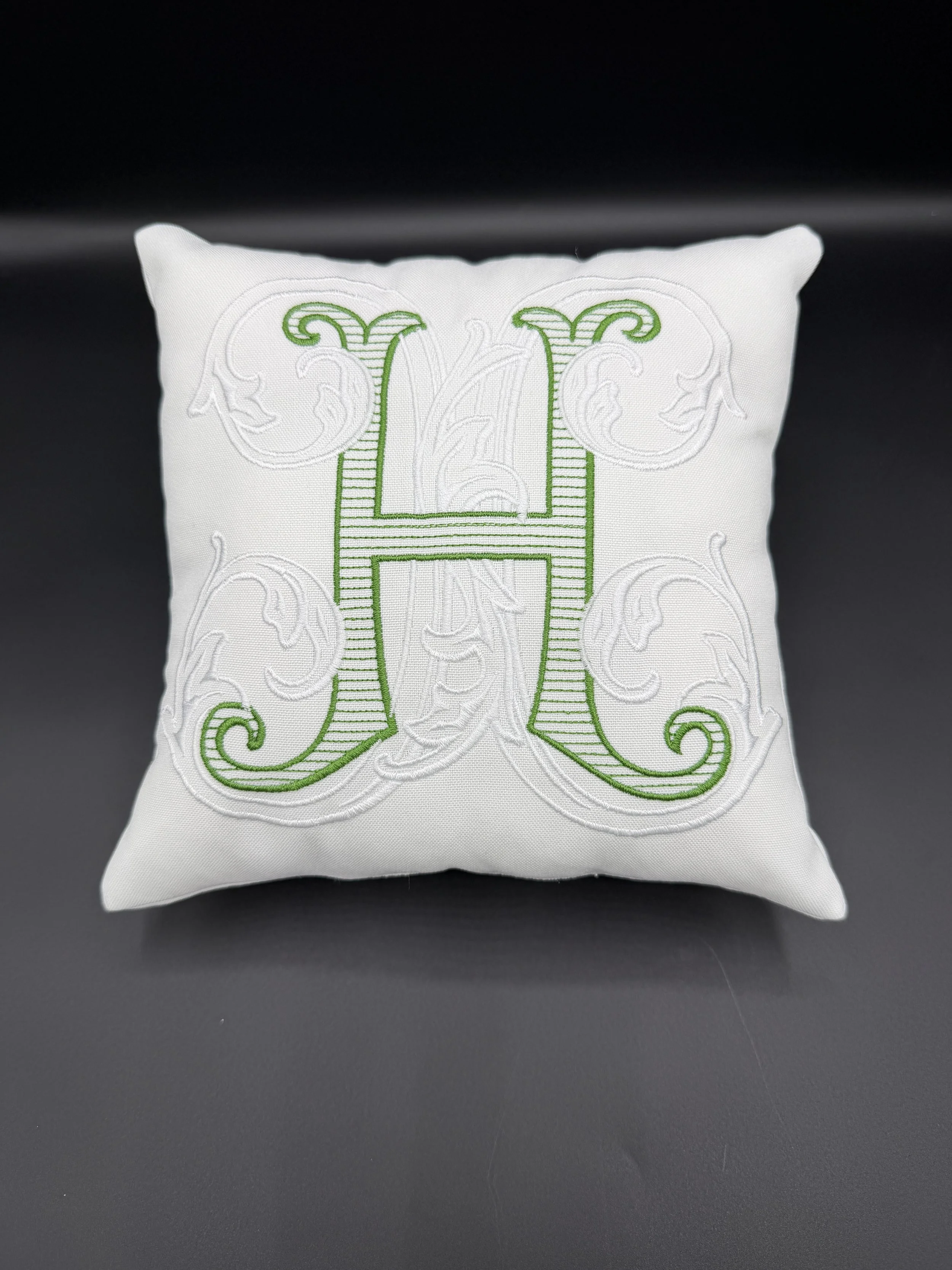 A white decorative pillow with green embroidery forming a monogram of the letter 'H' on a dark surface.