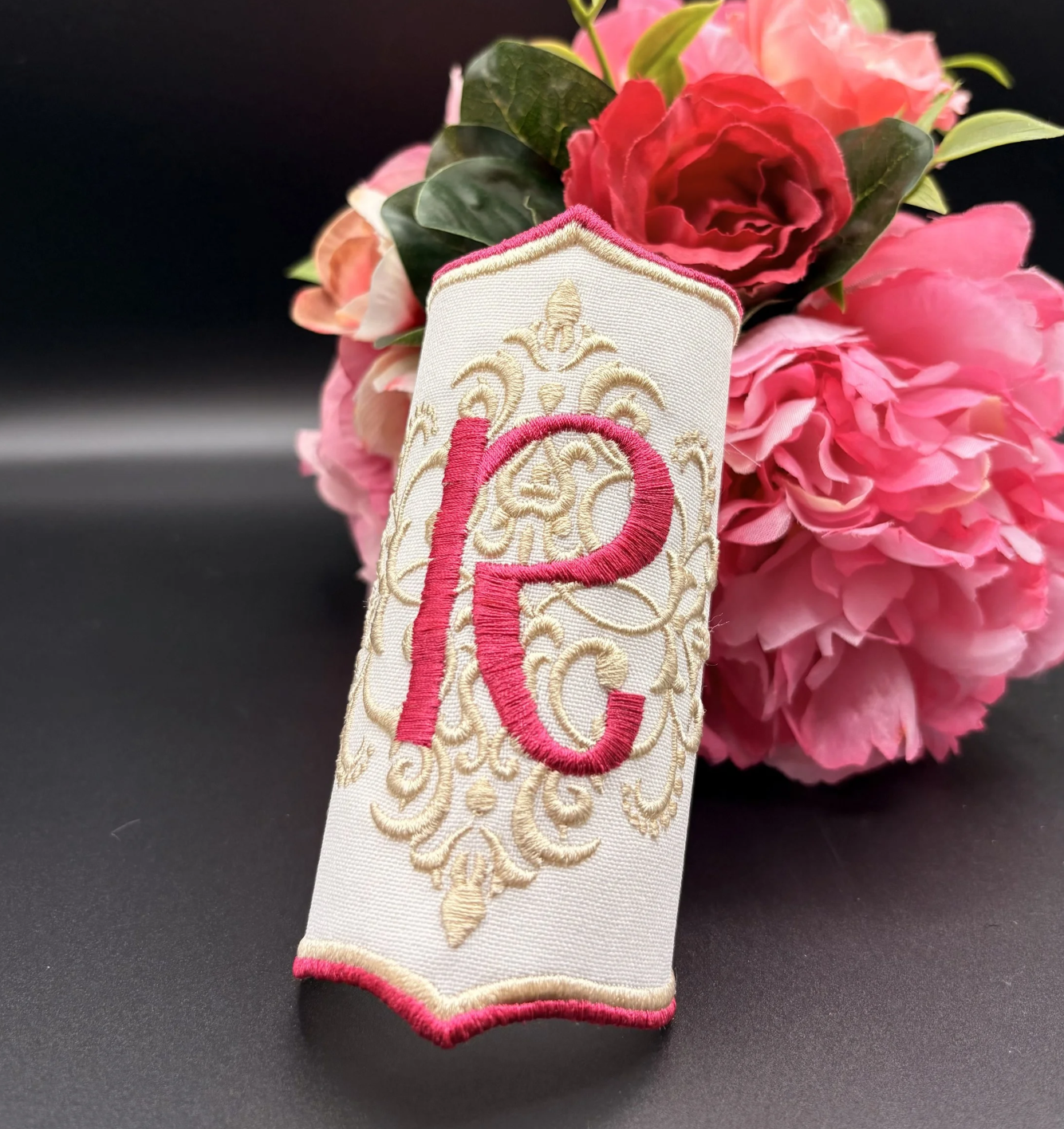 A decorative embroidered fabric piece with a pink and gold monogram 'P' and intricate gold embroidery, placed in front of pink and red flowers on a black background.