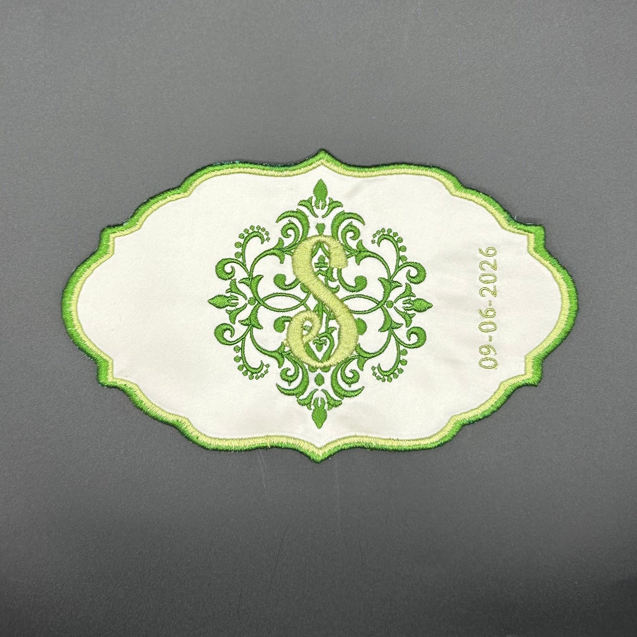 Close-up of an embroidered design on a white fabric with green thread, featuring a symmetrical ornate pattern against a gray background.