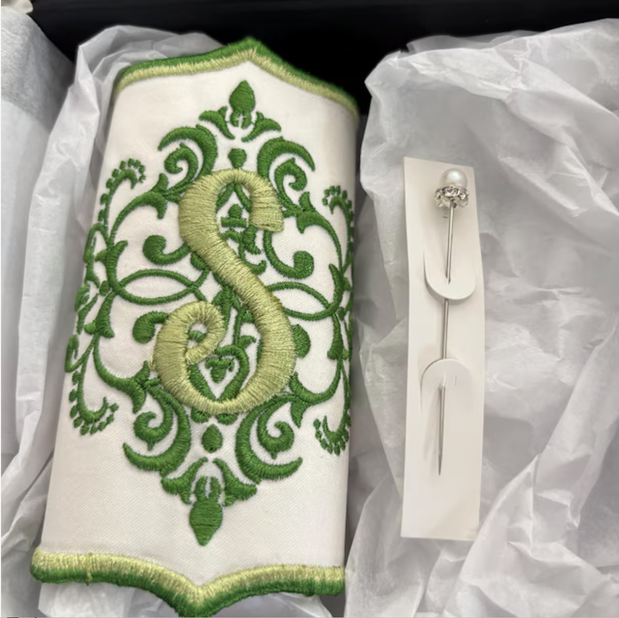 A decorative embroidery piece with green ornate swirl design, a large number six embroidered in gold, and green accents, paired with a pearl and rhinestone pin on a white background.