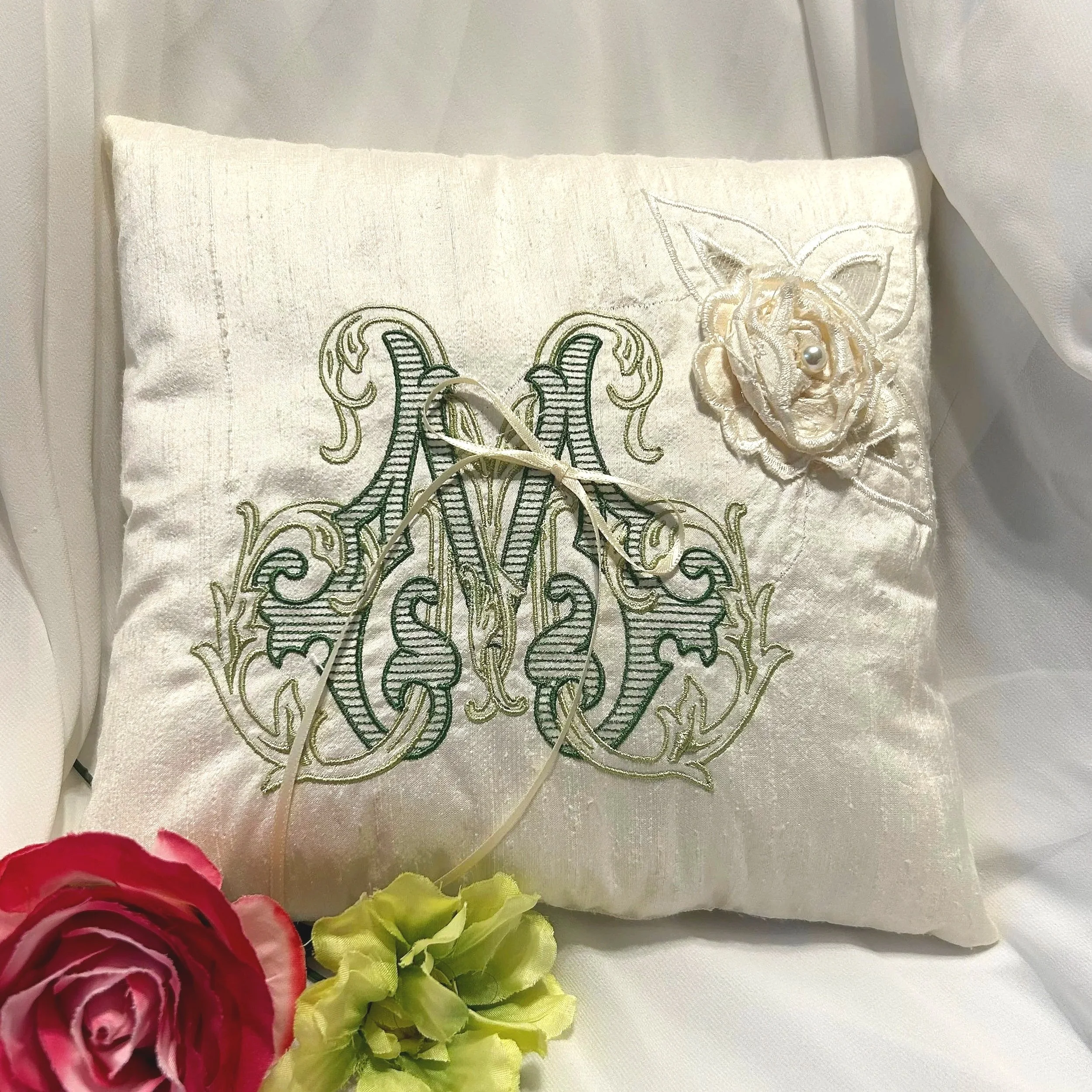 A decorative pillow with embroidered initials and a flower appliqué, surrounded by artificial flowers including red and green blooms, against a white fabric background.
