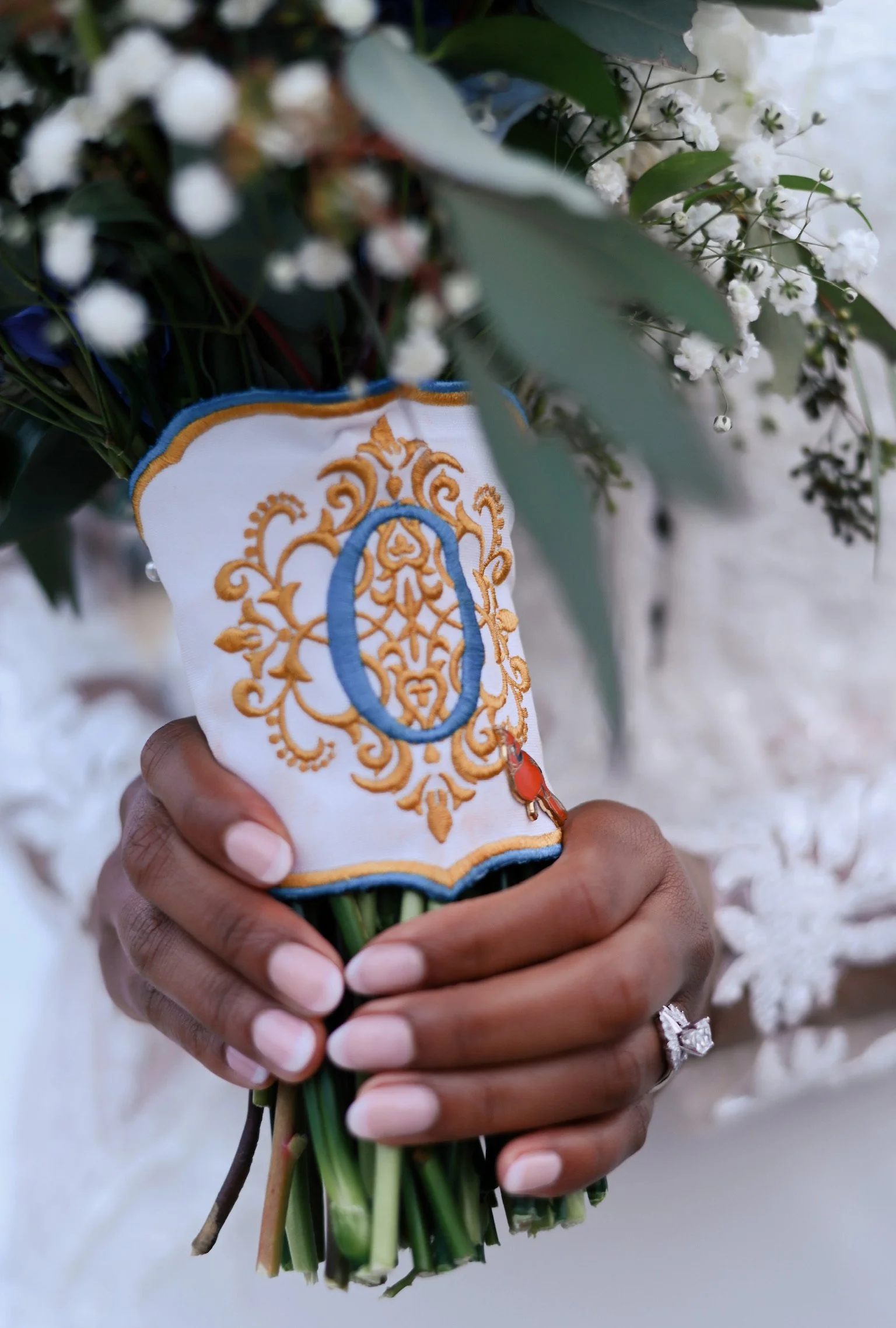 A close-up of a hand holding a bouquet of white flowers wrapped in a cloth with gold and blue embroidery.