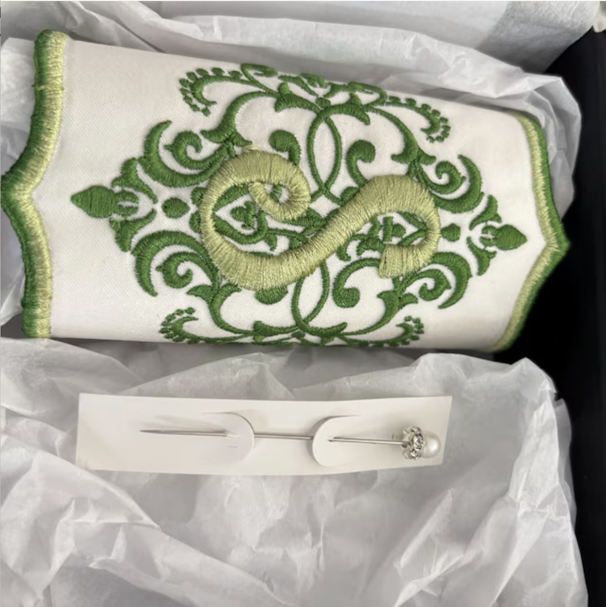 A white embroidered towel with green ornate patterns and a curled green design in the center, placed on white tissue paper, accompanied by a decorative pin with a pearl and rhinestone embellishment.