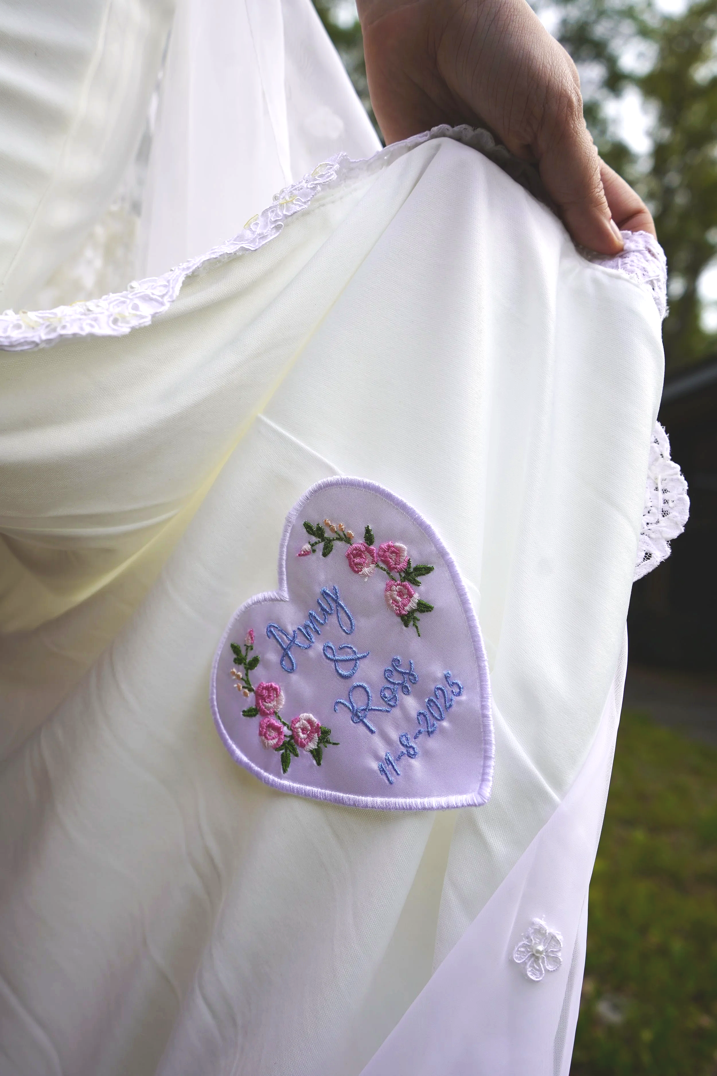 Close-up of a white wedding dress with a heart-shaped embroidered patch reading "June & Ross 11.8.2025" and floral embroidery, being gently held by a person's hand outside with blurred trees in the background.