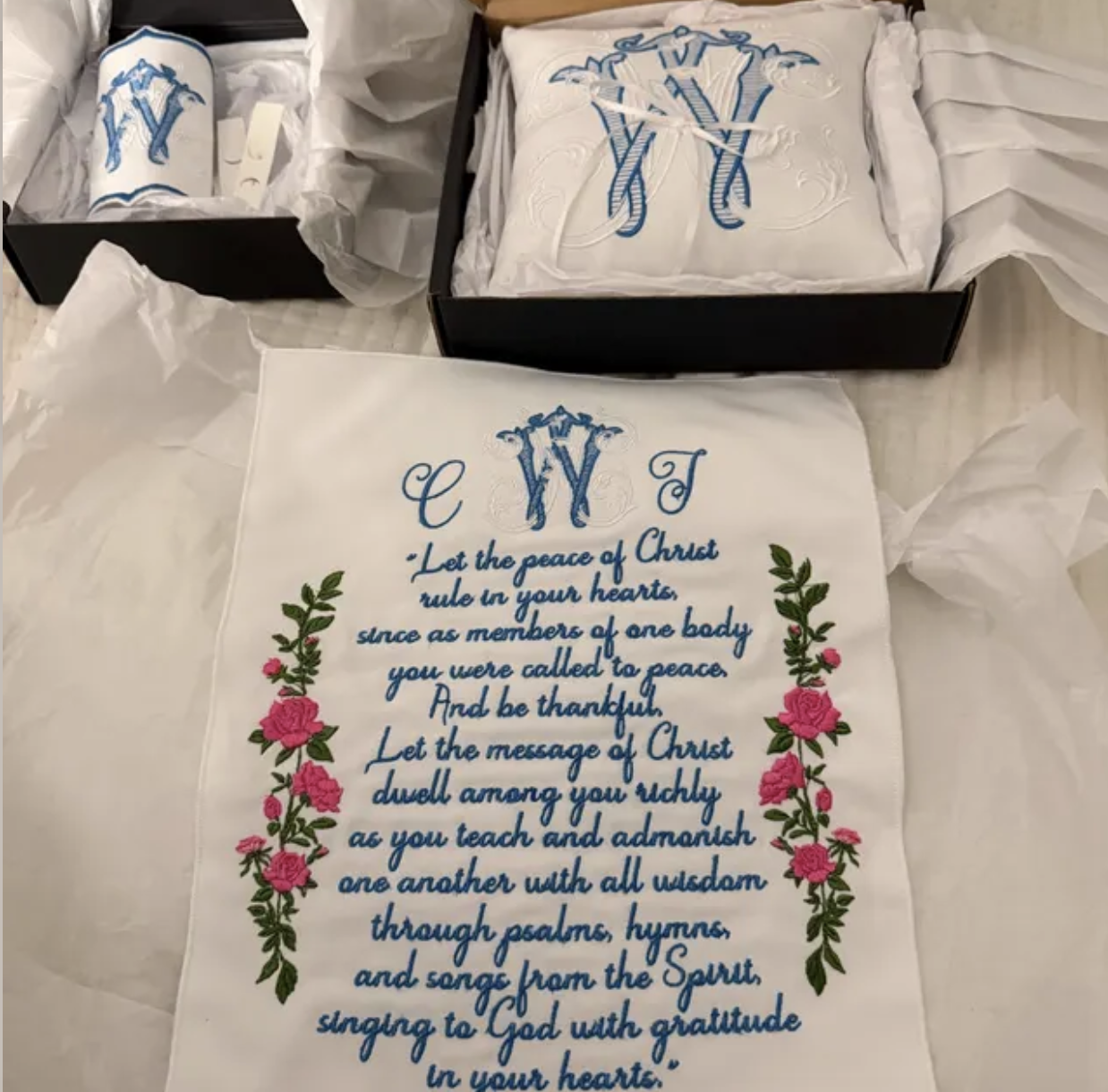A white embroidered cloth with a Christian prayer and floral decoration, placed on a table with tissue paper. There are also boxed items with embroidered monograms in the background, featuring the initials V and Y intertwined.