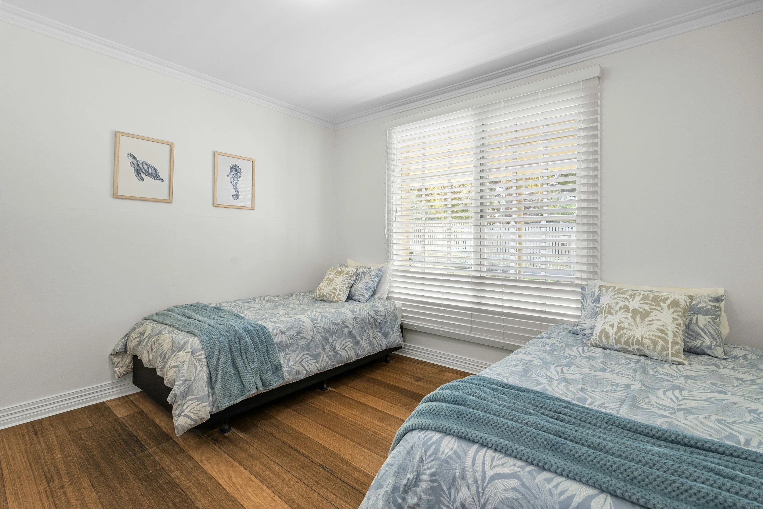 The image shows a bedroom with two beds covered in light blue bedding with floral patterns, a large window with white blinds allowing natural light, framed artwork of sea creatures on a white wall, and hardwood flooring.