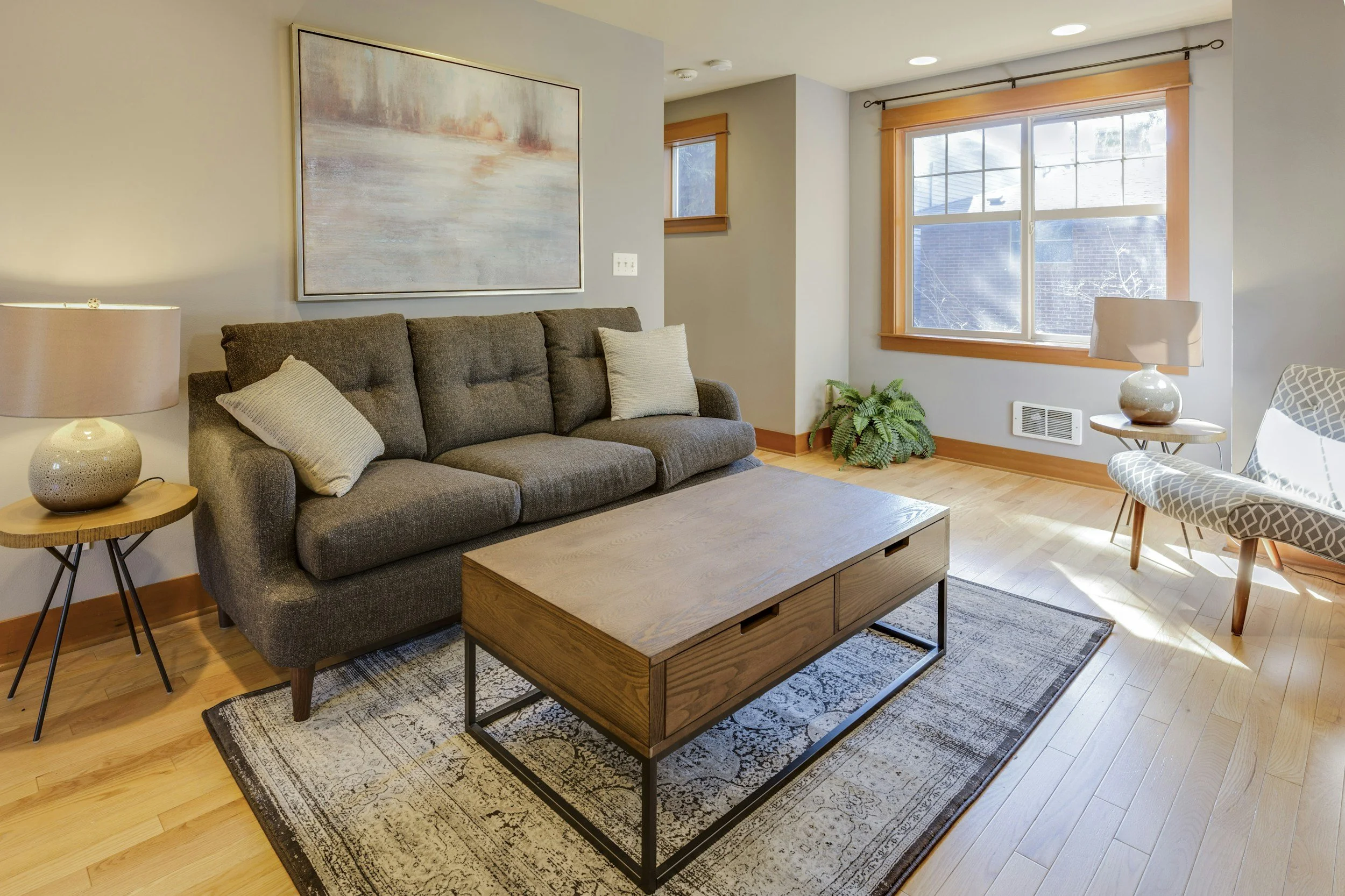 A living room with a dark gray sofa, light wood coffee table, area rug, two lamps, a woven chair, framed artwork, and large windows with wood trim allowing natural light