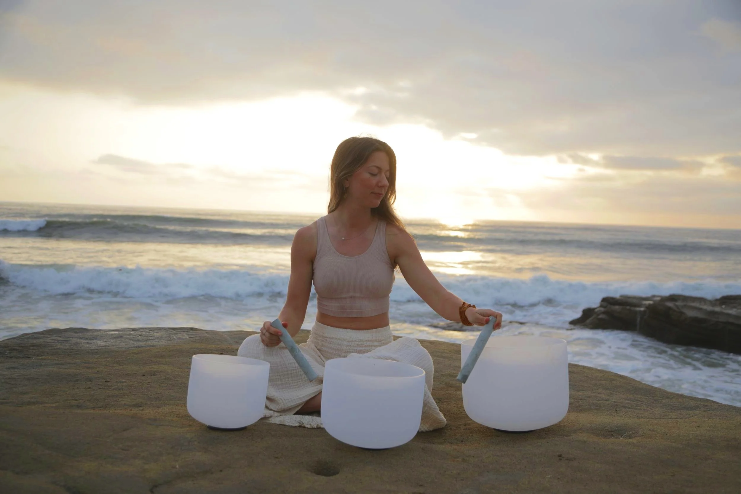 yoga teacher and sound healer sitting on a rocky shoreline, playing crystal singing bowls at sunset with ocean waves in the background san diego.