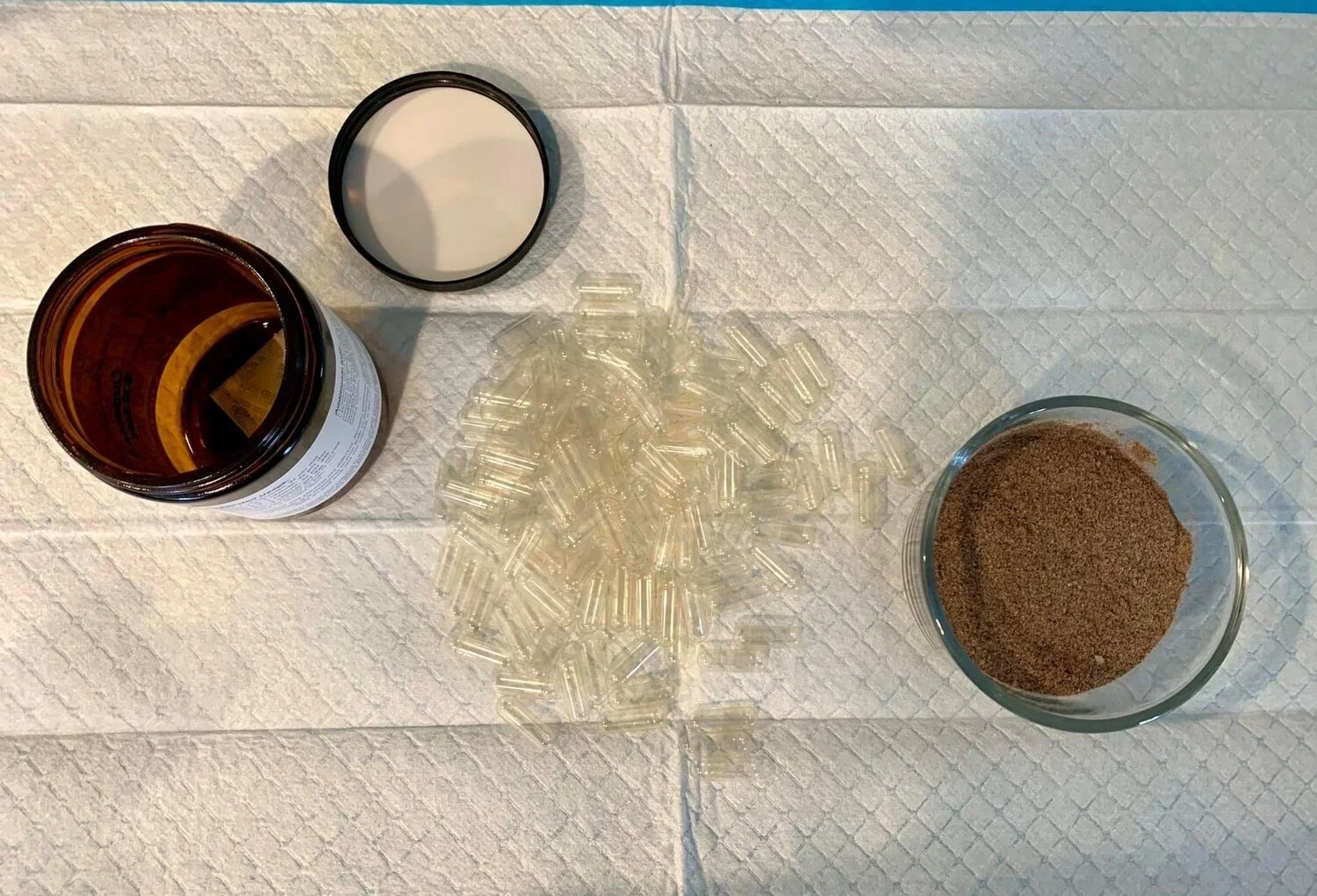 Brown glass jar with lid, empty gelatin capsules, and ground powder in a bowl on a white textured surface.