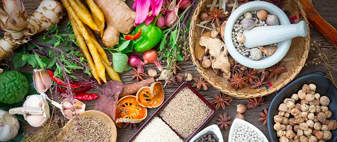Assorted herbs and spices including ginger, turmeric, peppers, onions, orange slices, and seeds on a wooden surface.